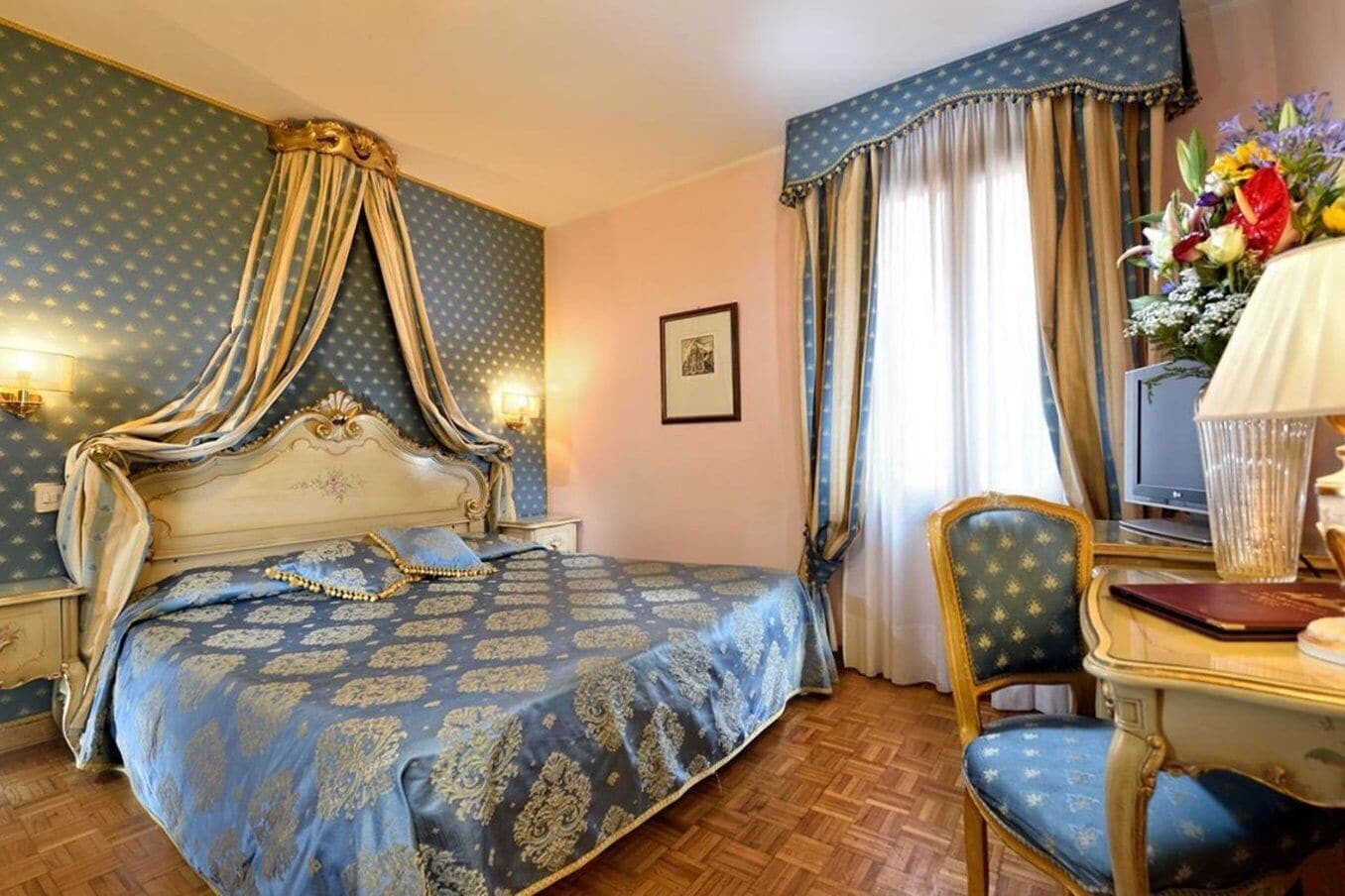 A bedroom with a four-poster bed, a desk with a chair, a television, and a vase of flowers.