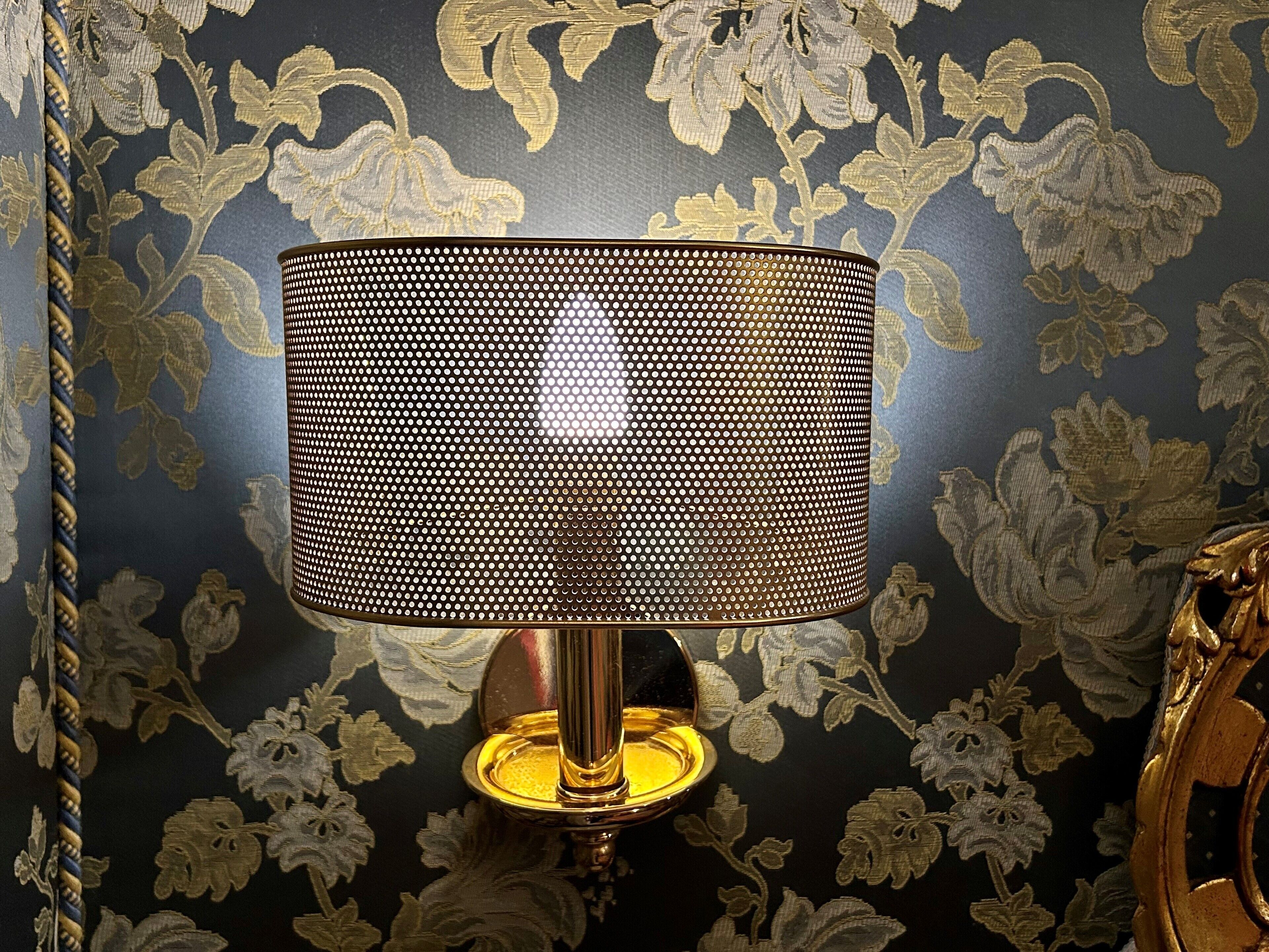 A lamp with a perforated shade on a stand, set against a floral wallpaper background.