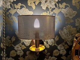Una lámpara con pantalla calada sobre un pedestal, frente a un fondo de papel tapiz con motivos florales.
