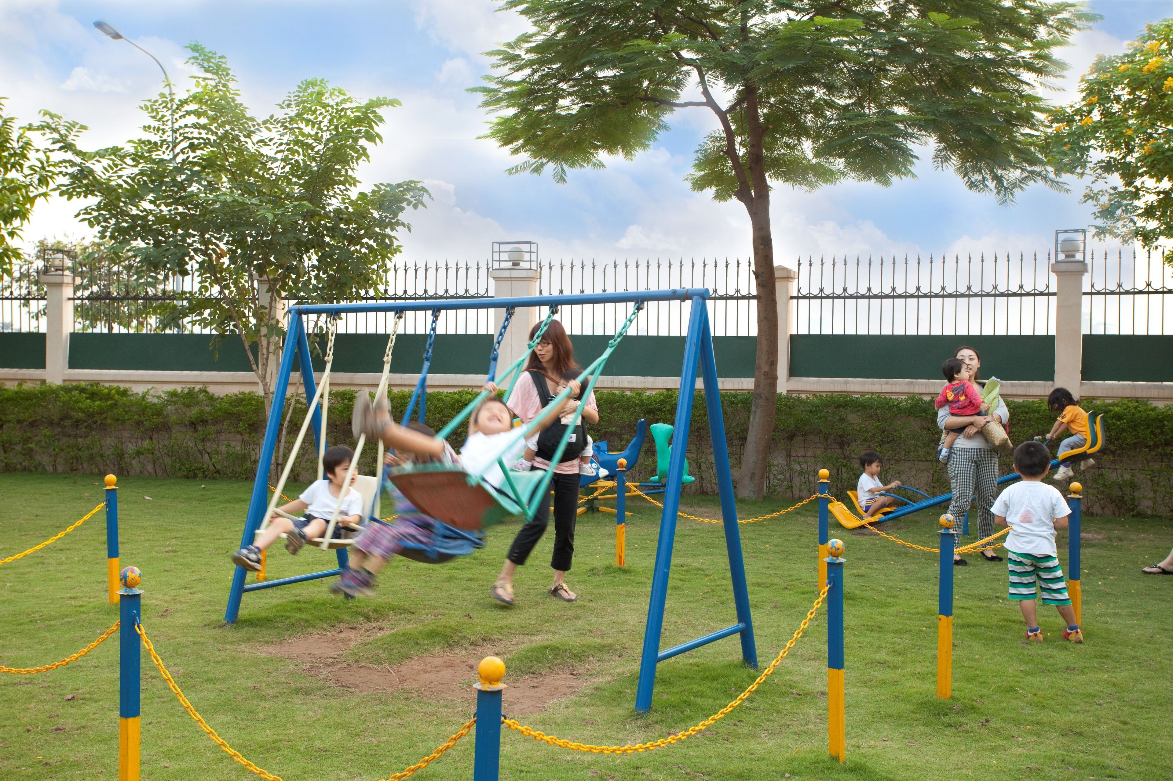 A playground with swings and a fenced area, surrounded by greenery and a white fence.