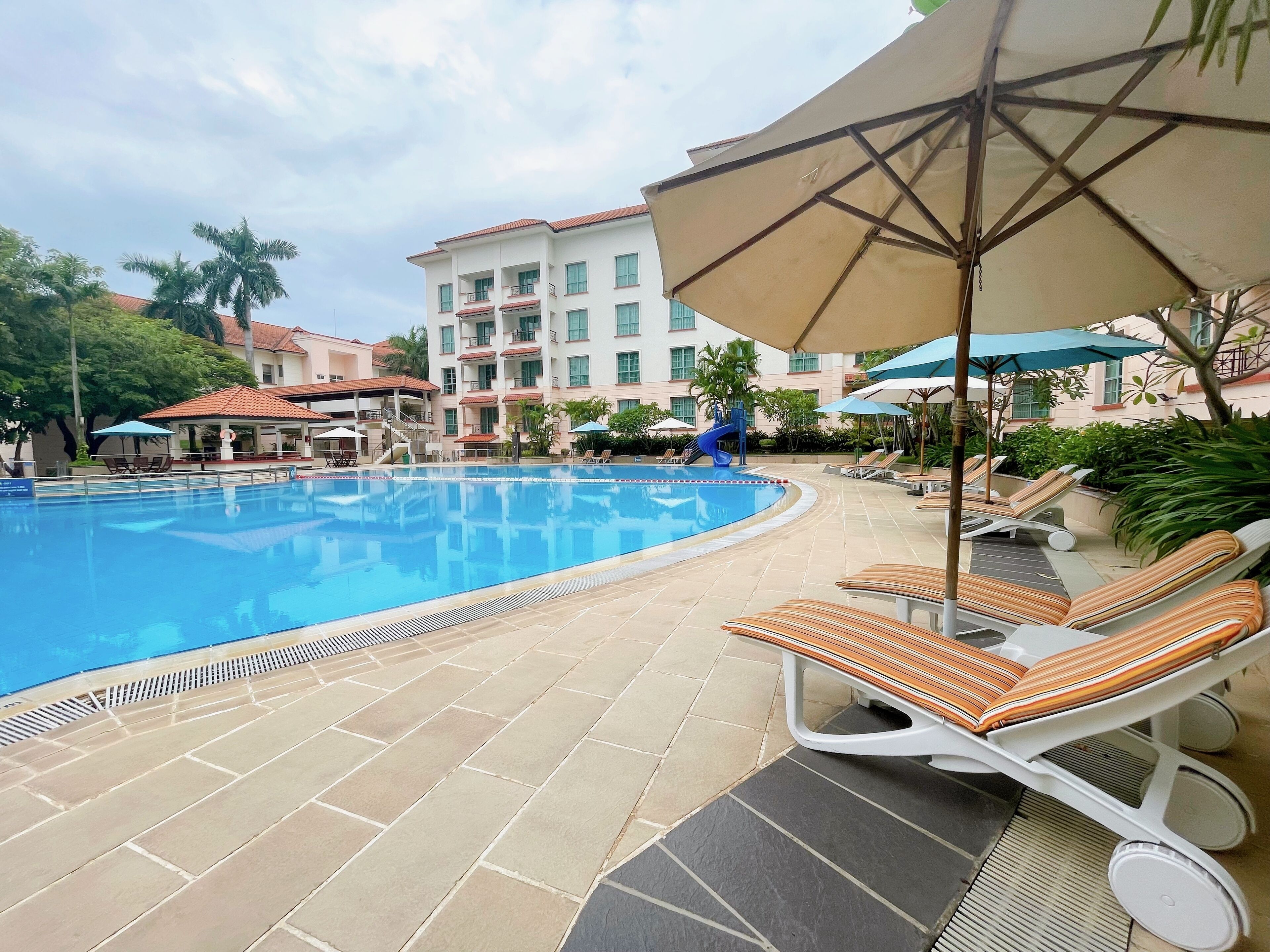 A swimming pool with lounge chairs and umbrellas, surrounded by a building with multiple balconies and a covered area.