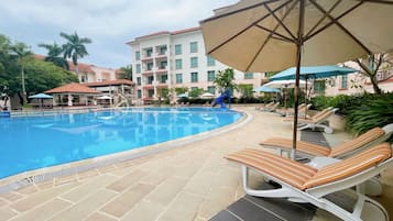 A swimming pool with lounge chairs and umbrellas, surrounded by a building with multiple balconies and a covered area.