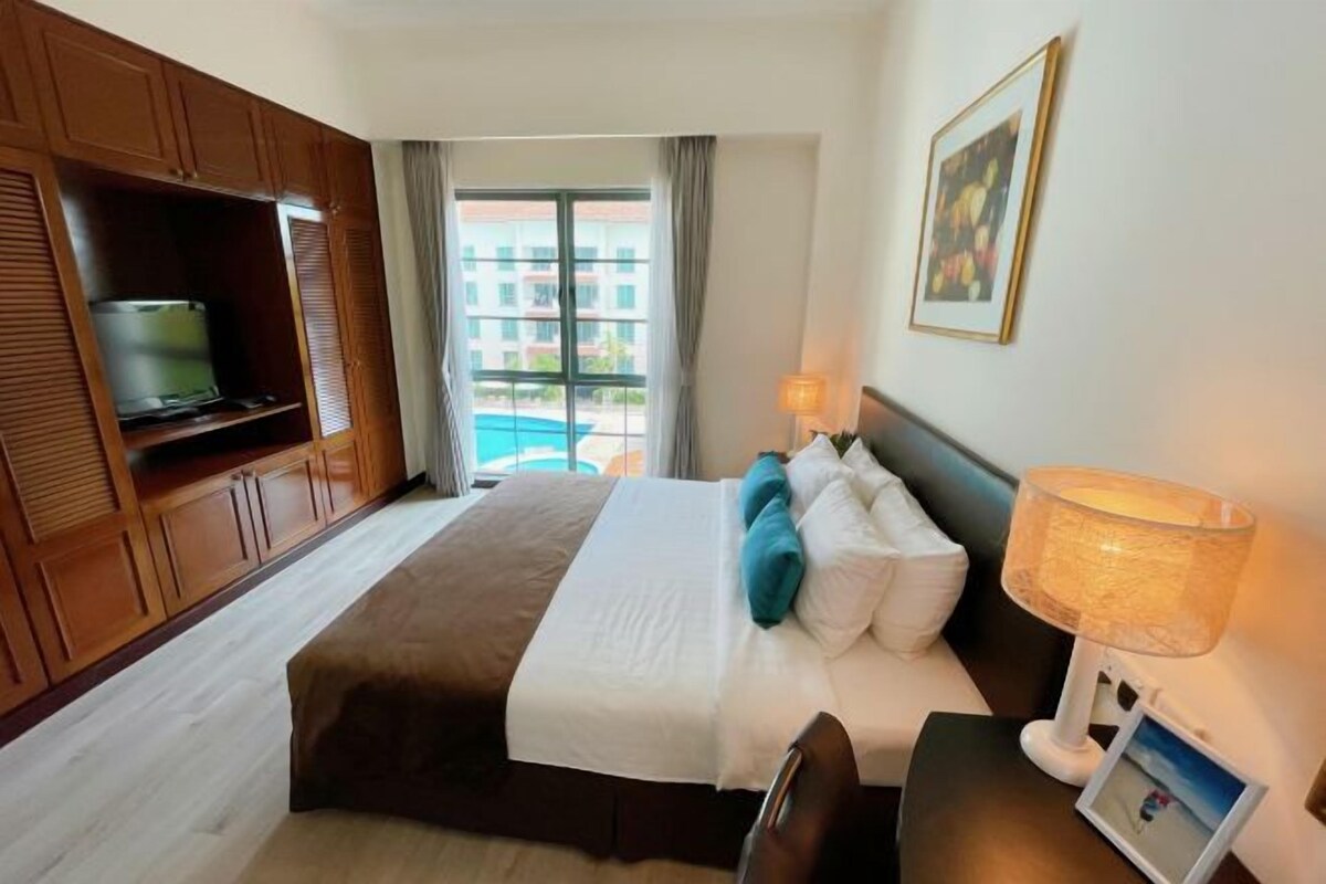 A hotel room with a bed, a TV on a wooden cabinet, a lamp, and a window with a view of buildings.