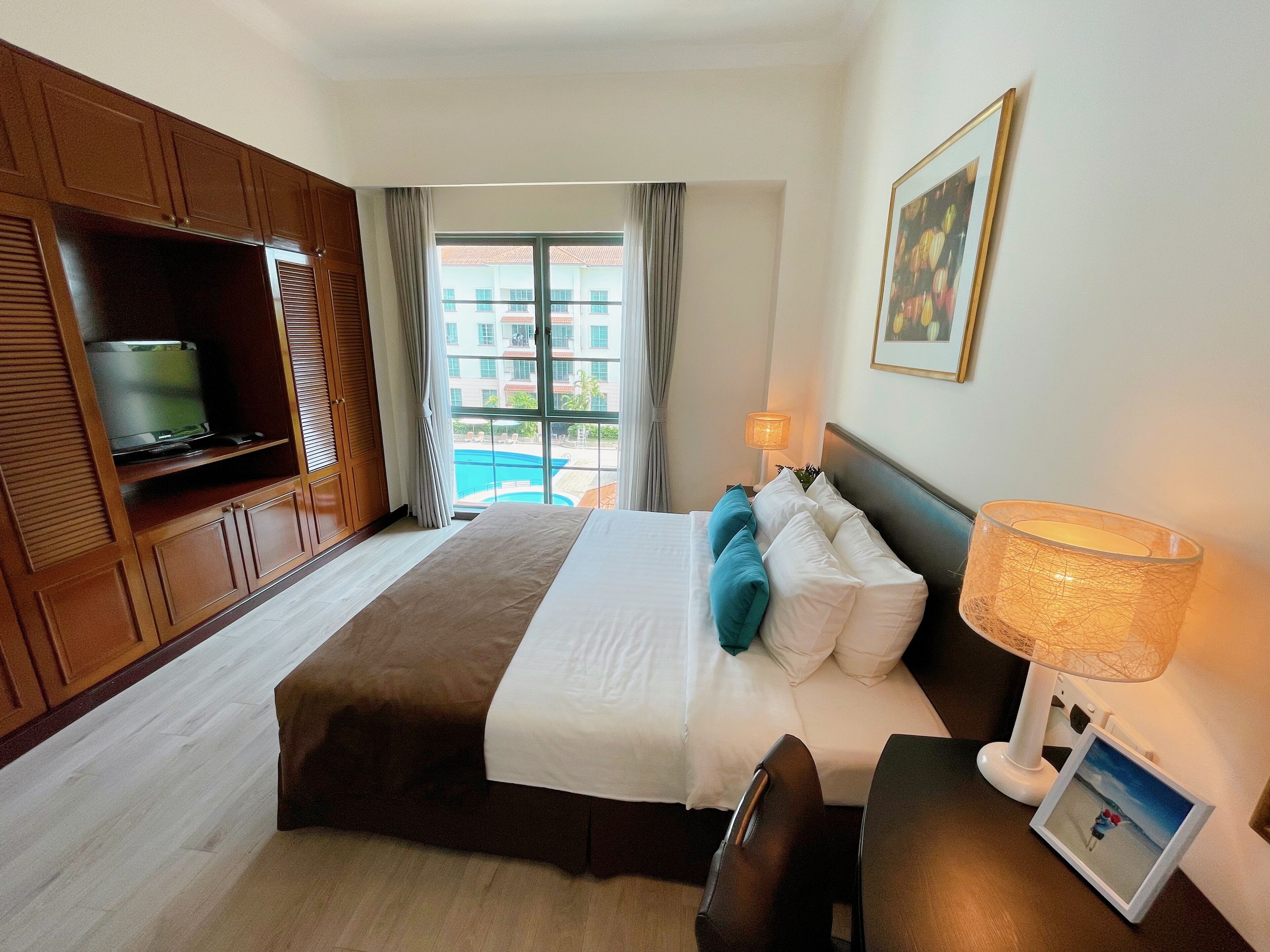A hotel room with a bed, a TV, a lamp, a picture frame, and a view of a swimming pool.
