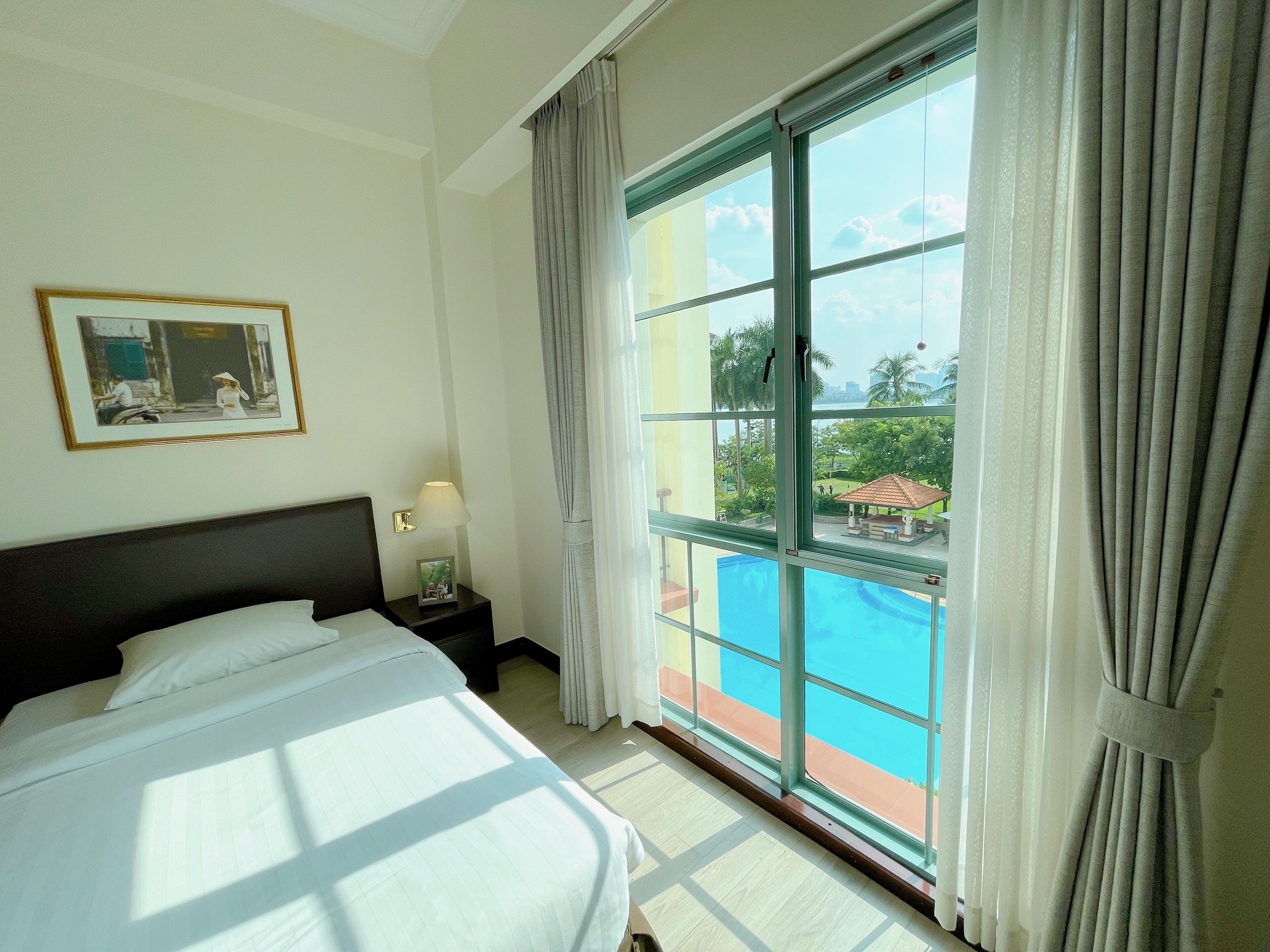 A hotel room with a large bed, a nightstand with a lamp, a framed picture on the wall, and a view of a swimming pool and outdoor area.