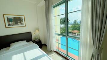 A hotel room with a large bed, a nightstand with a lamp, a framed picture on the wall, and a view of a swimming pool and outdoor area.
