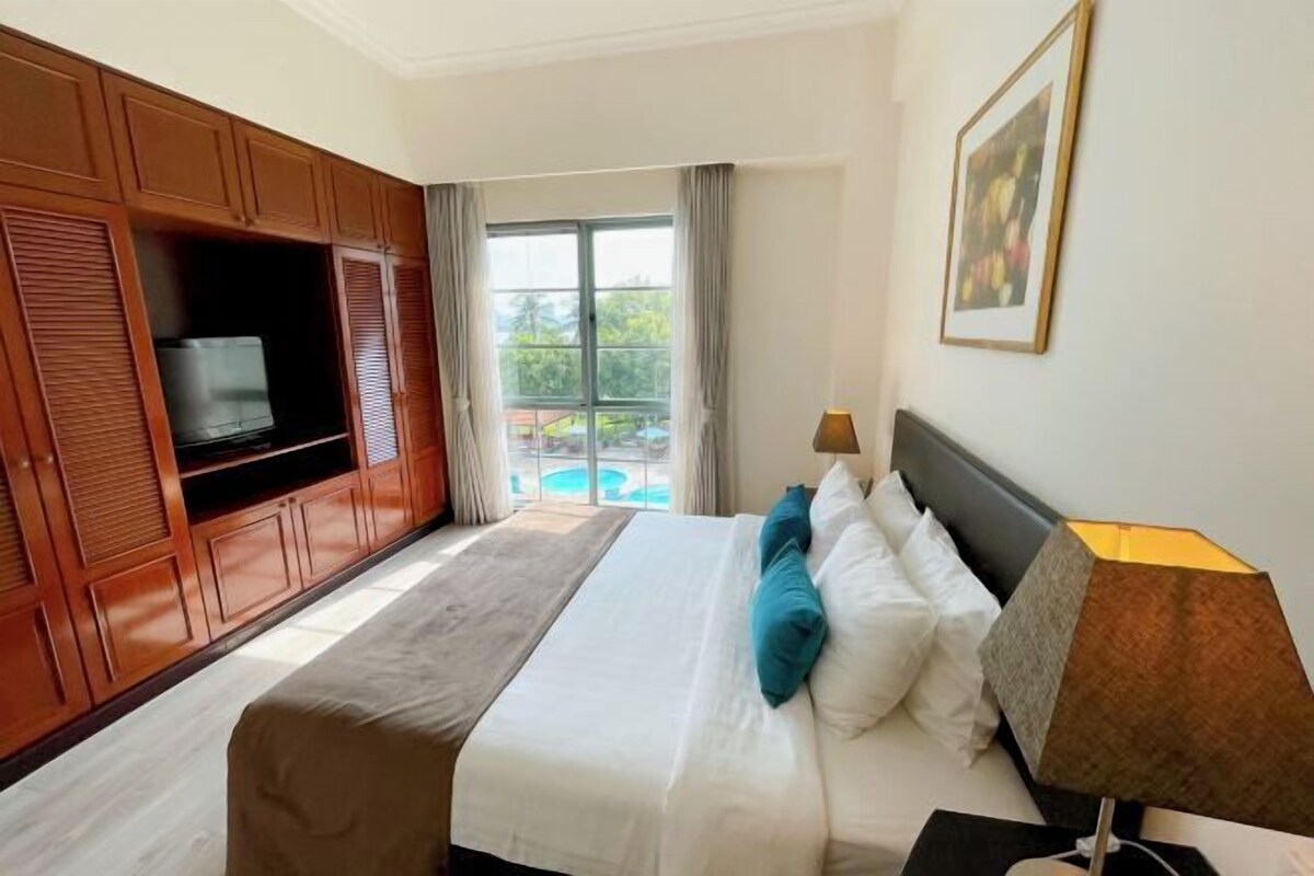 A hotel room with a bed, a TV, a lamp, and a view of a pool.