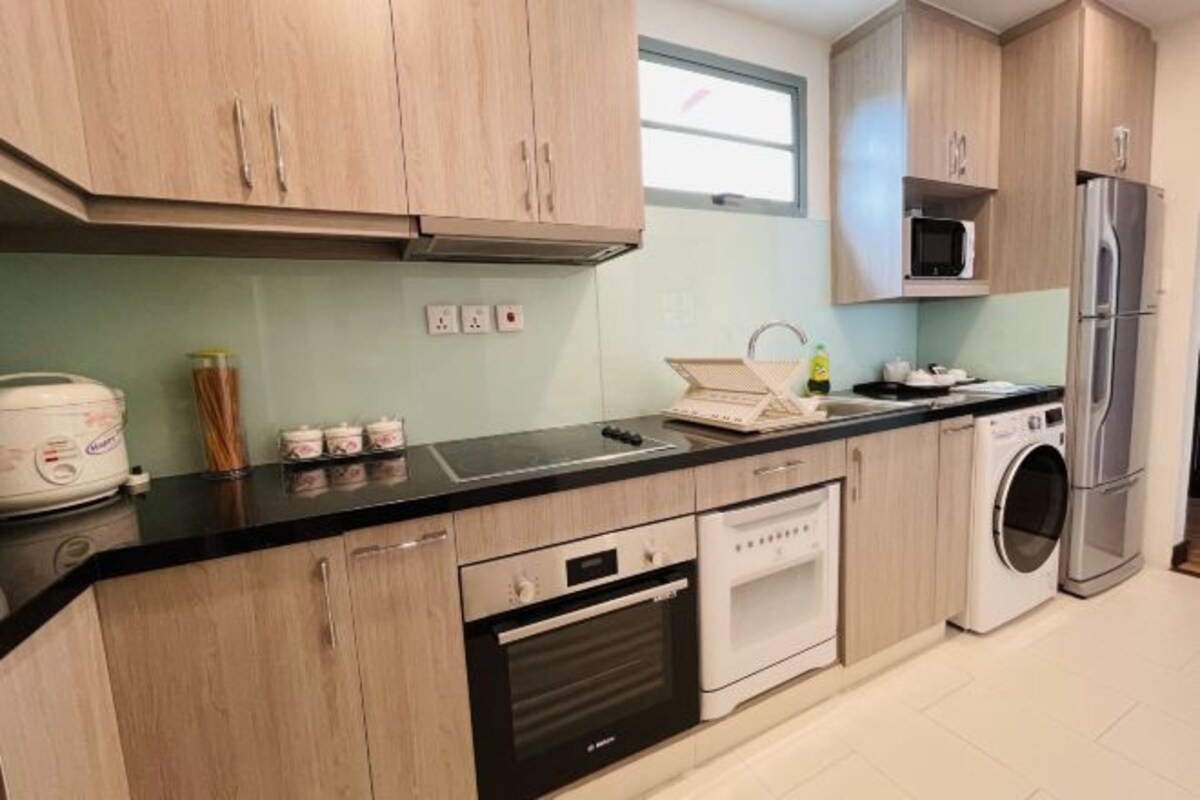 A modern kitchen with wooden cabinets, a black countertop, a built-in oven, a microwave, and a washing machine.