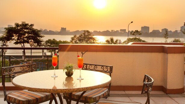 Mesa redonda con sillas y dos bebidas en una terraza con vista a un cuerpo de agua y el horizonte de una ciudad al fondo.