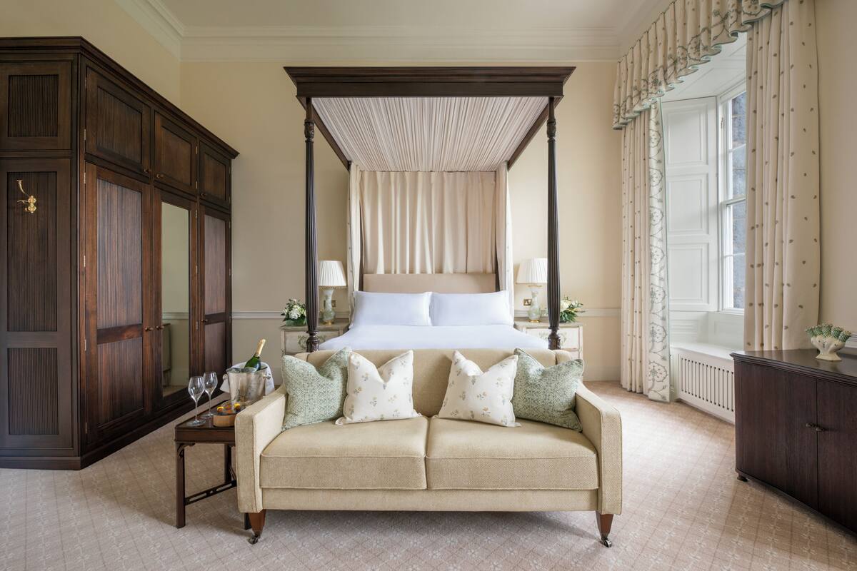 Een hemelbed met houten hoofdeinde, een beige bank met bloemenkussens, een nachtkastje met lamp en een grote, donkerhouten garderobe.