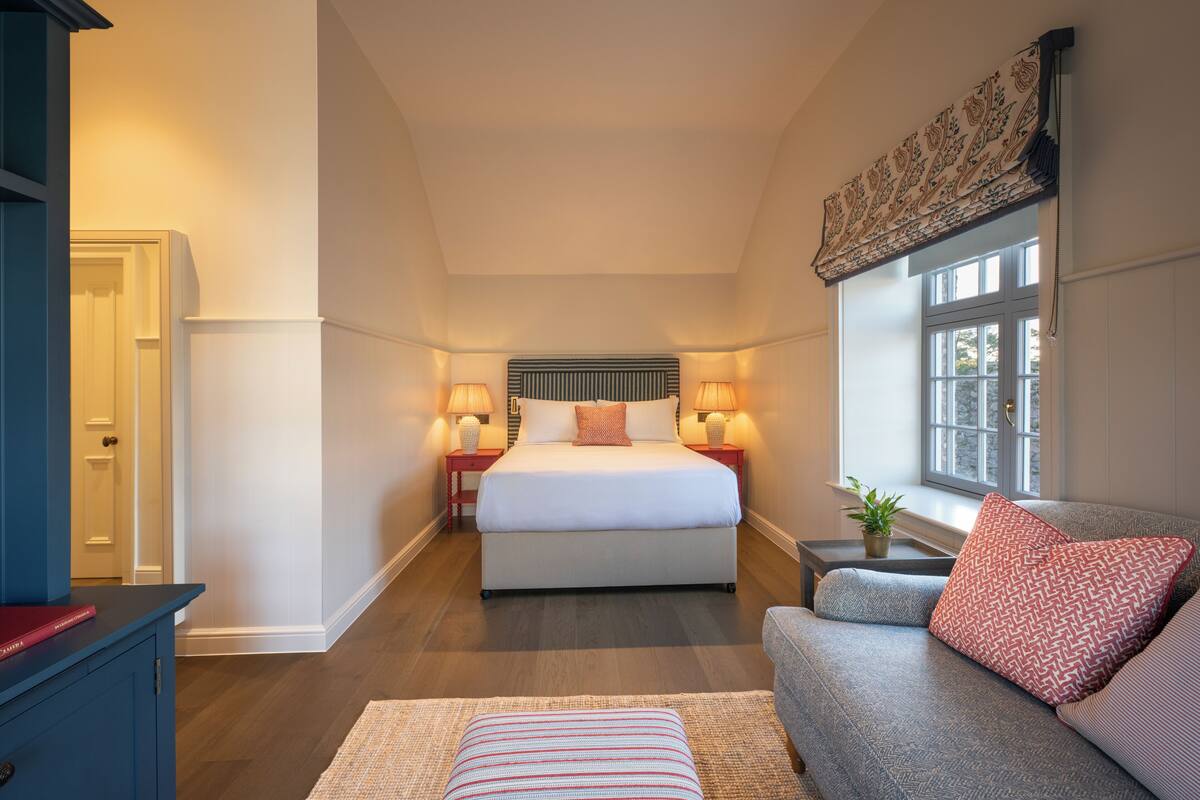 Een slaapkamer met een groot bed, bedlampen, een raam met uitzicht en een bank met een gekleurd kussen.