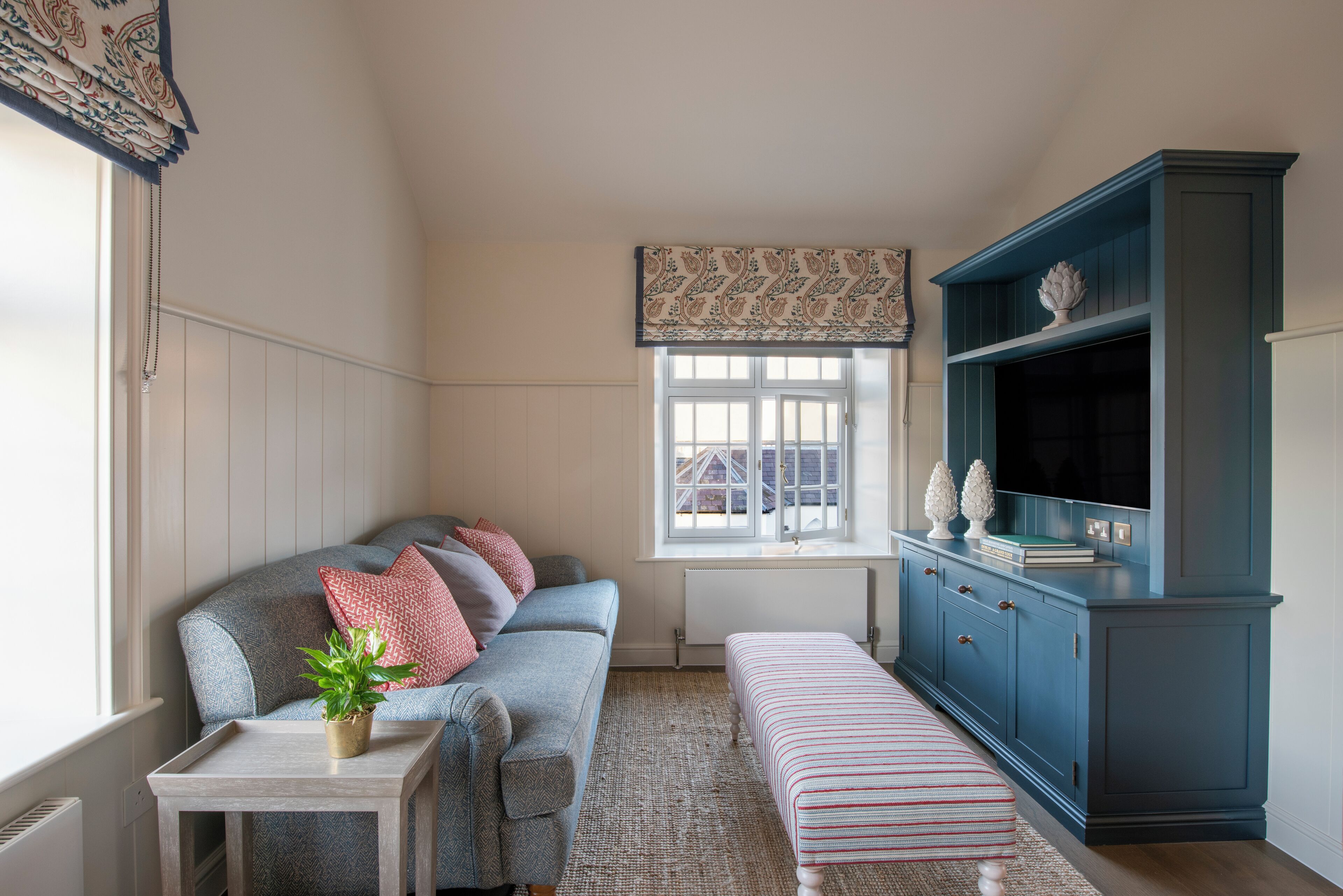 A cozy living room with a grey sofa, a small table with a plant, a striped bench, and a blue cabinet with a television.