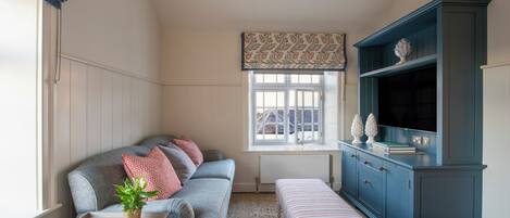 Una sala de estar acogedora con un sofá gris, una mesita con una planta, un banco a rayas y un mueble azul con un televisor.
