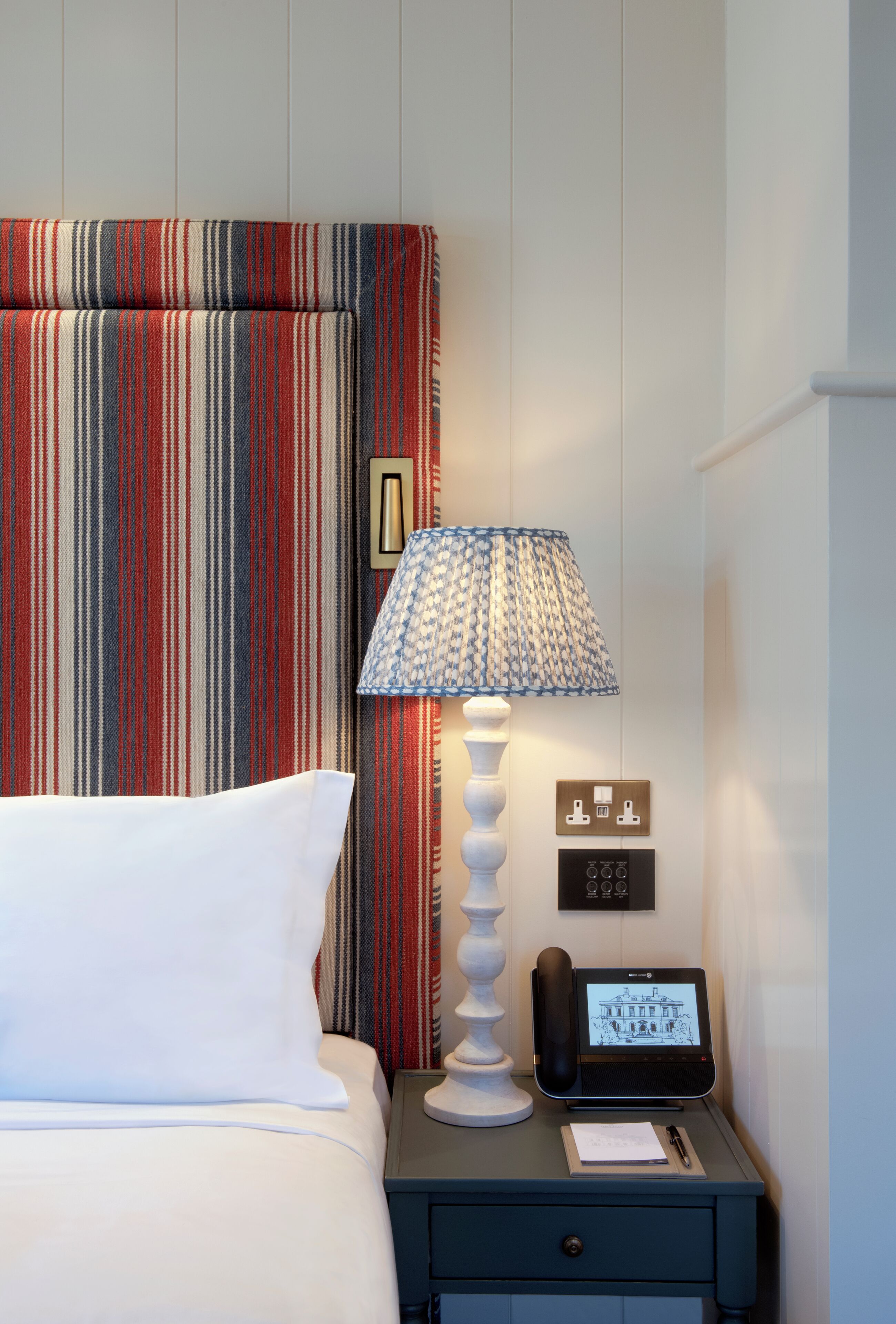 A hotel room with a striped headboard, a bedside lamp, a bedside table with a phone and a notepad, and a bed with white bedding.