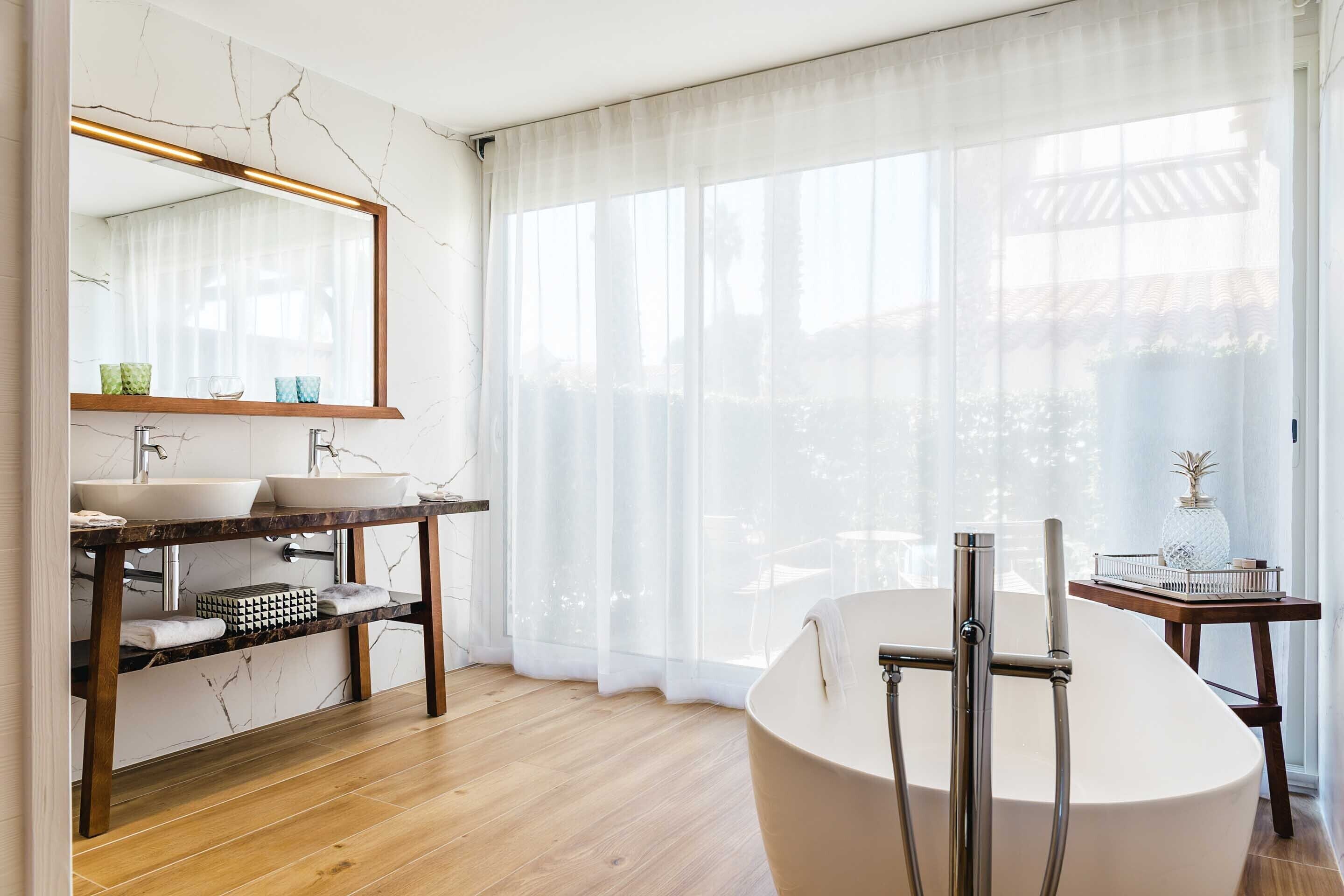 Une salle de bain moderne avec un meuble-lavabo en marbre, une baignoire sur pieds et une grande fenêtre agrémentée de rideaux légers.