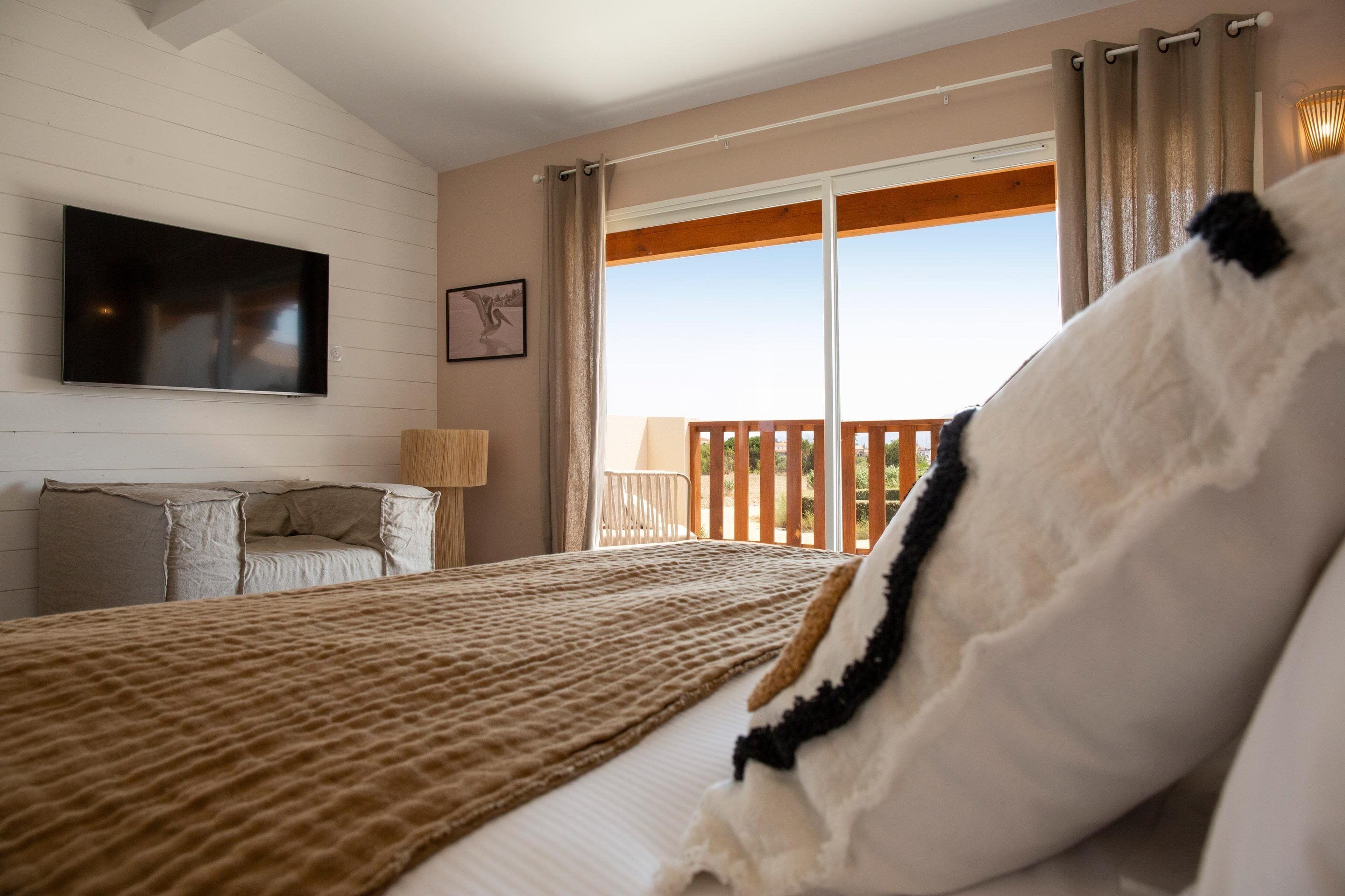 A bedroom with a large bed, a flat-screen TV mounted on the wall, and a balcony with a wooden railing.