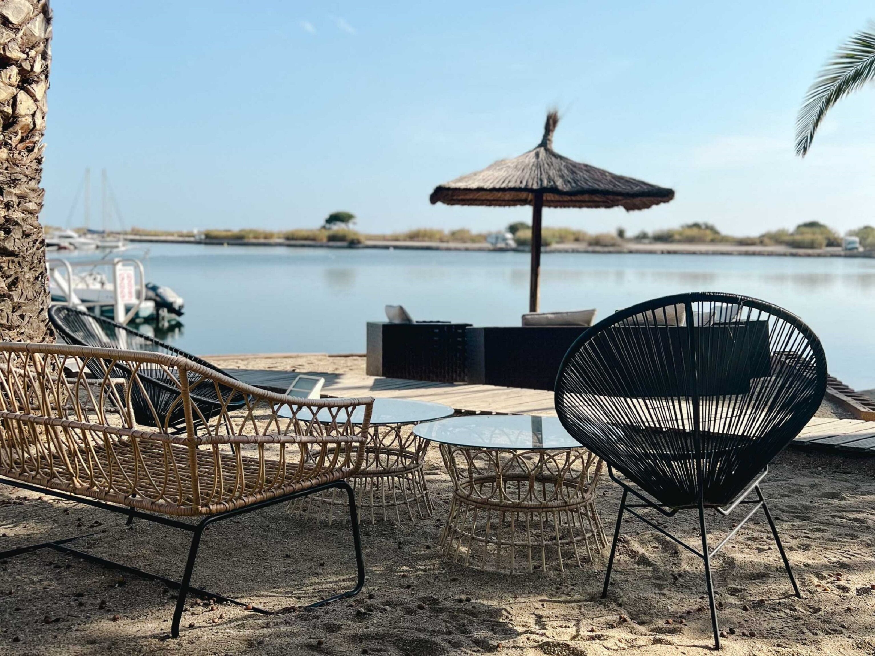 Un área de estar frente a la playa con sillas de mimbre y una mesa redonda, una sombrilla de paja y vista al agua y a los barcos al fondo.