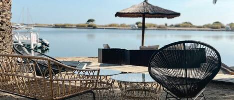 Un área de estar frente a la playa con sillas de mimbre y una mesa redonda, una sombrilla de paja y vista al agua y a los barcos al fondo.