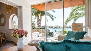 A modern room with a large sliding glass door leading to a balcony with a view of palm trees and a body of water. There is a round wooden table with a vase of flowers, a blue sofa, and a wicker chair. - Relais et Châteaux Thalasso & Spa Ile de la Lagune (Saint-Cyprien)