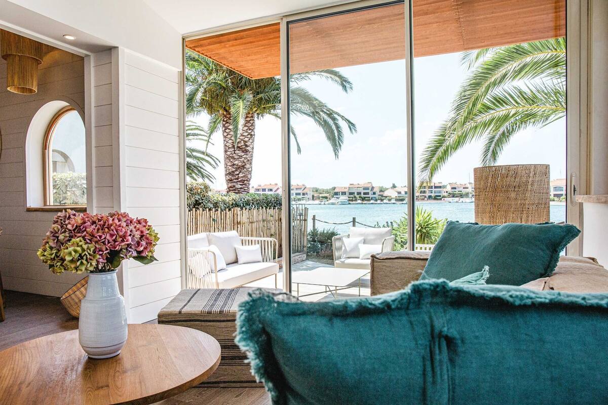 Ein modernes Zimmer mit einer großen Schiebetür, die auf einen Balkon mit Blick auf Palmen und ein Gewässer führt. Es gibt einen runden Holztisch mit Blumen in einer Vase, ein blaues Sofa und einen Korbstuhl.