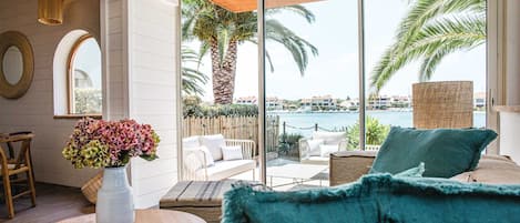 Habitación moderna con un amplio ventanal corredero que da a un balcón con vistas a palmeras y una masa de agua. Se observa una mesa redonda de madera con un florero, un sofá azul y una silla de mimbre.