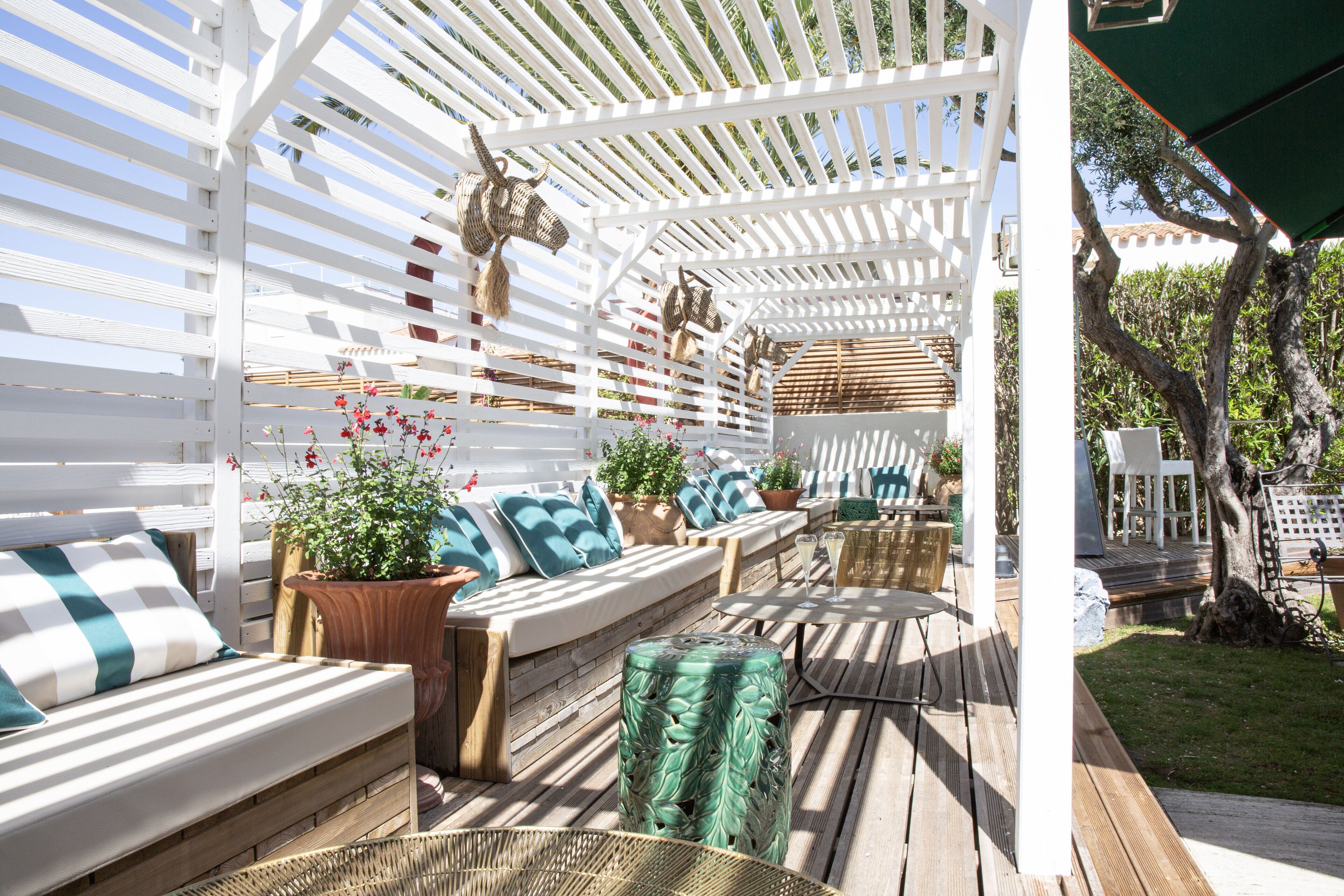 Un área de estar al aire libre techada, con bancos y cojines de madera, rodeada de plantas en macetas y una estructura de pérgola.