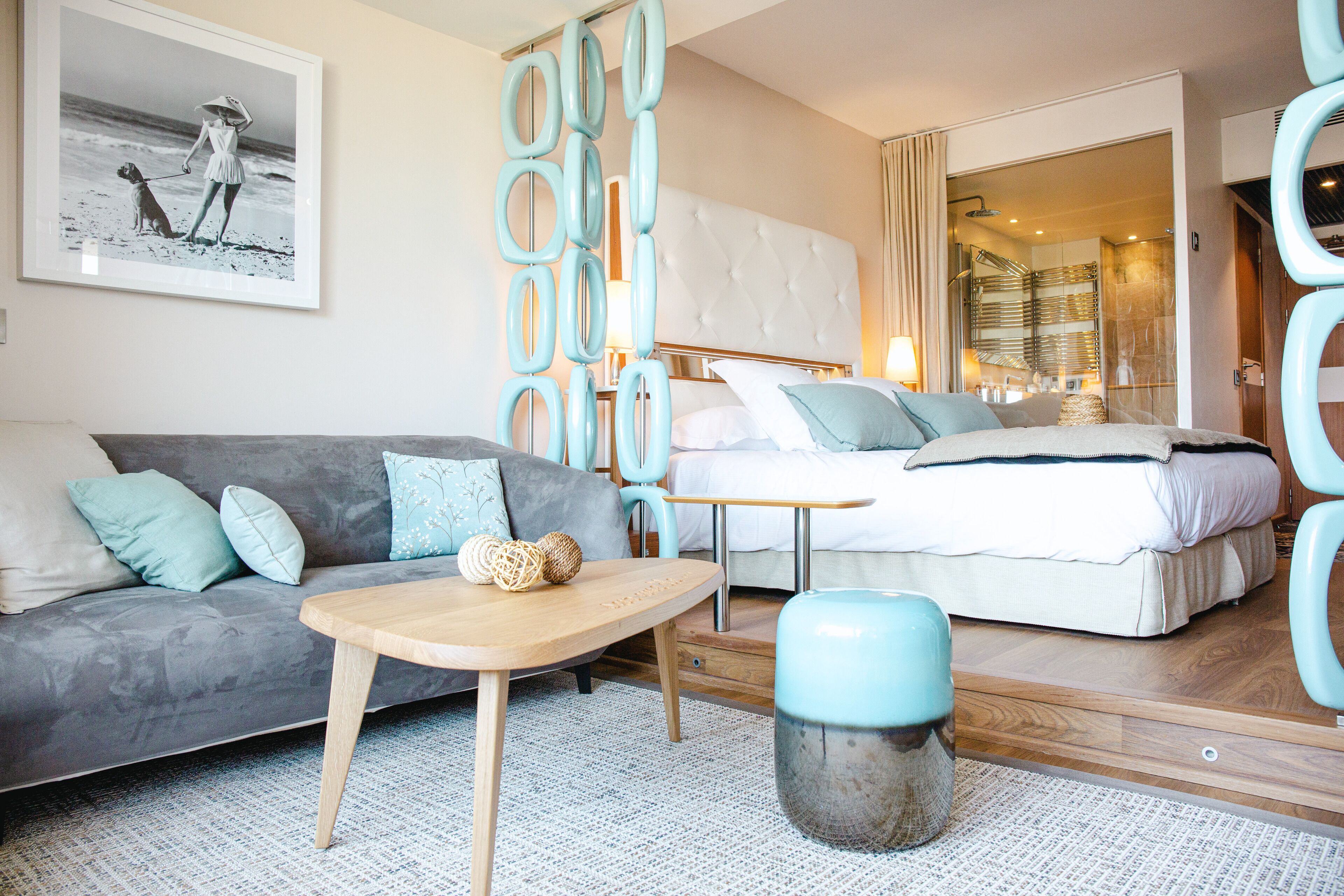 Una habitación de hotel moderna con un sofá gris, una mesa de madera, una cama con ropa de cama blanca y un baño visible a través de una puerta abierta.