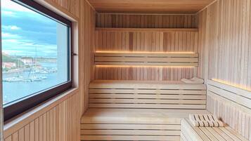 A sauna with wooden walls and a bench, a window with a view of boats and buildings, and a tiled floor.