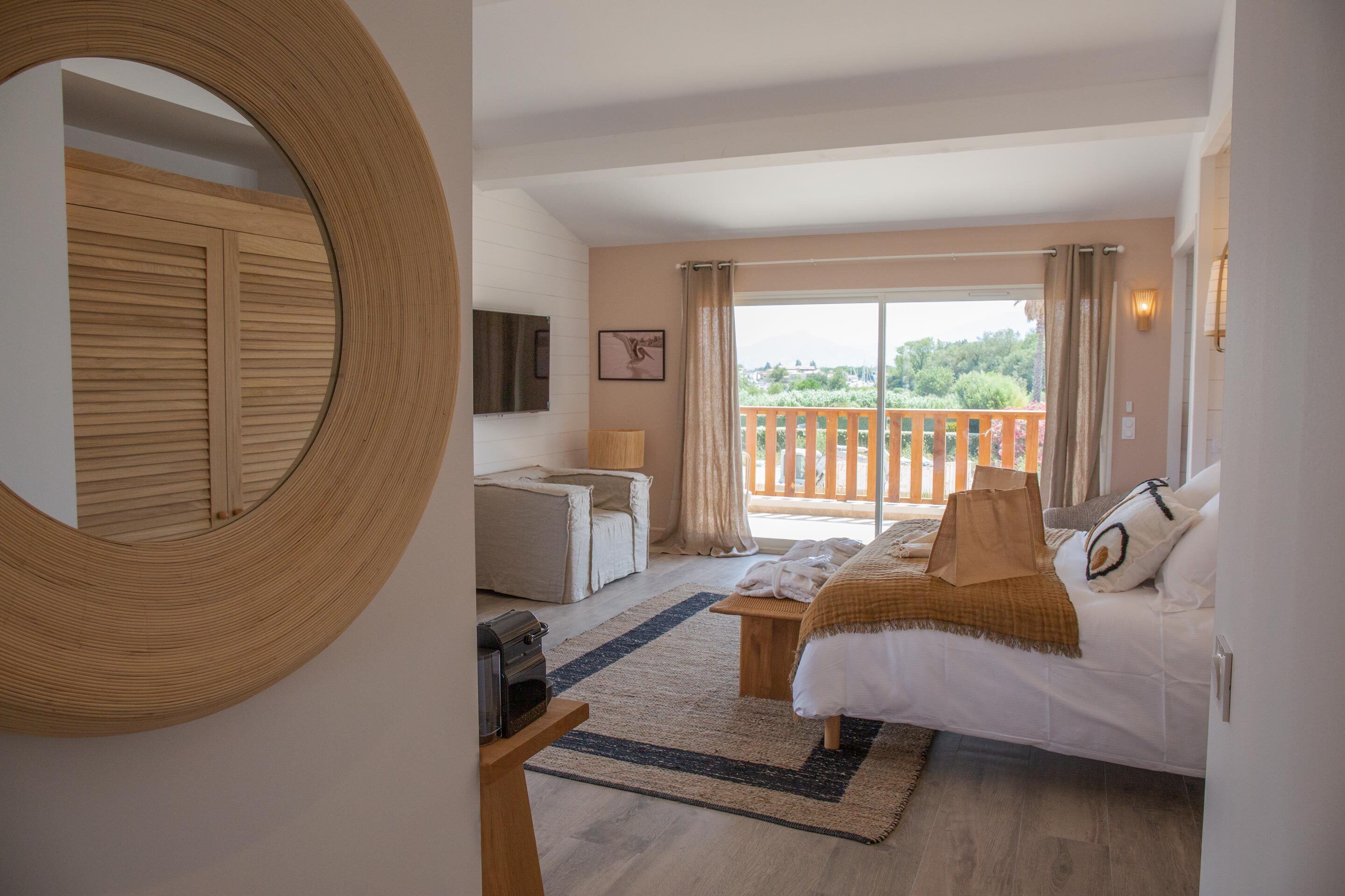 A bedroom with a bed, a desk, a chair, a large mirror, and a balcony with a view of greenery.