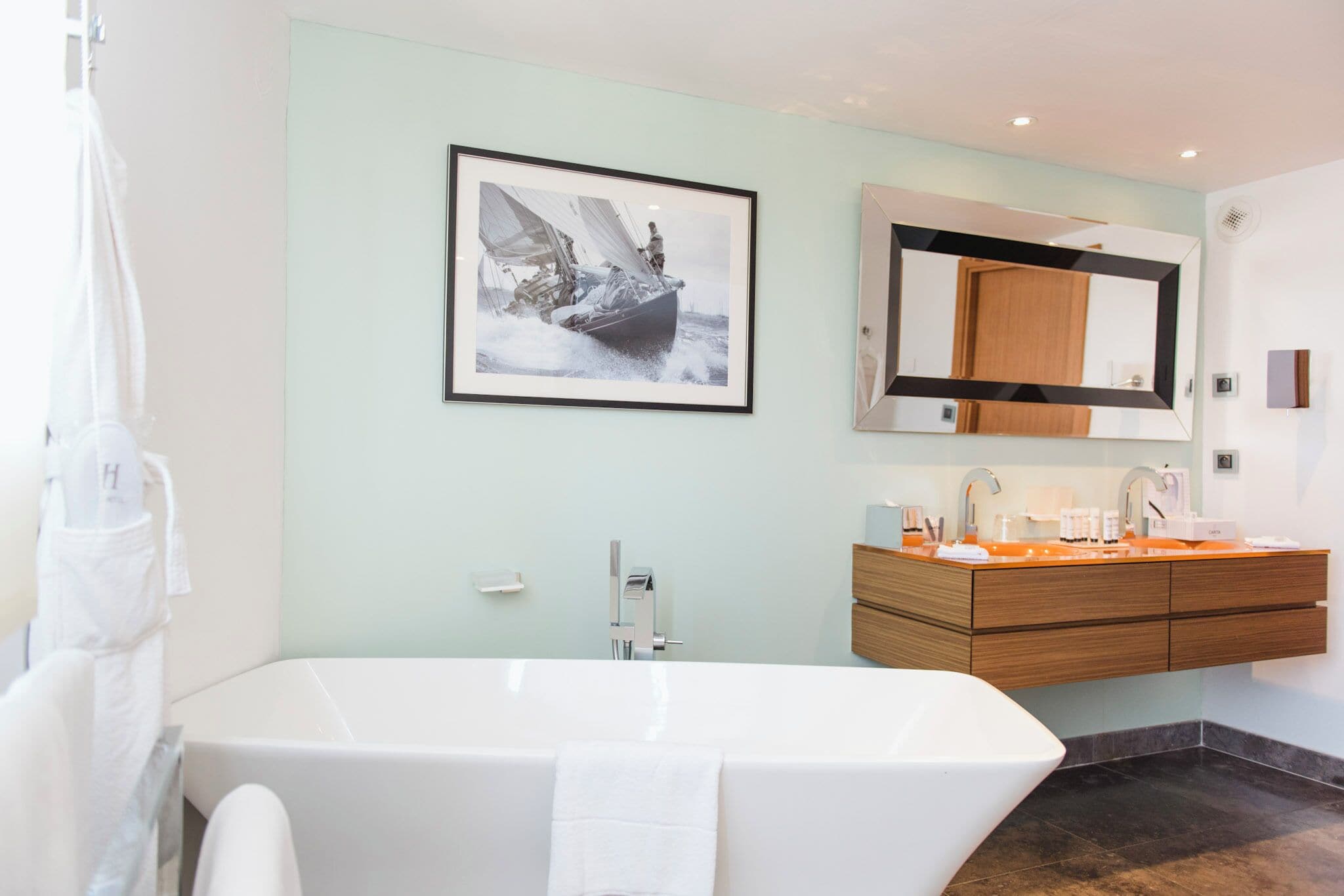 A modern bathroom with a freestanding bathtub, a large framed photograph of a sailboat, and a wooden vanity with a mirror.