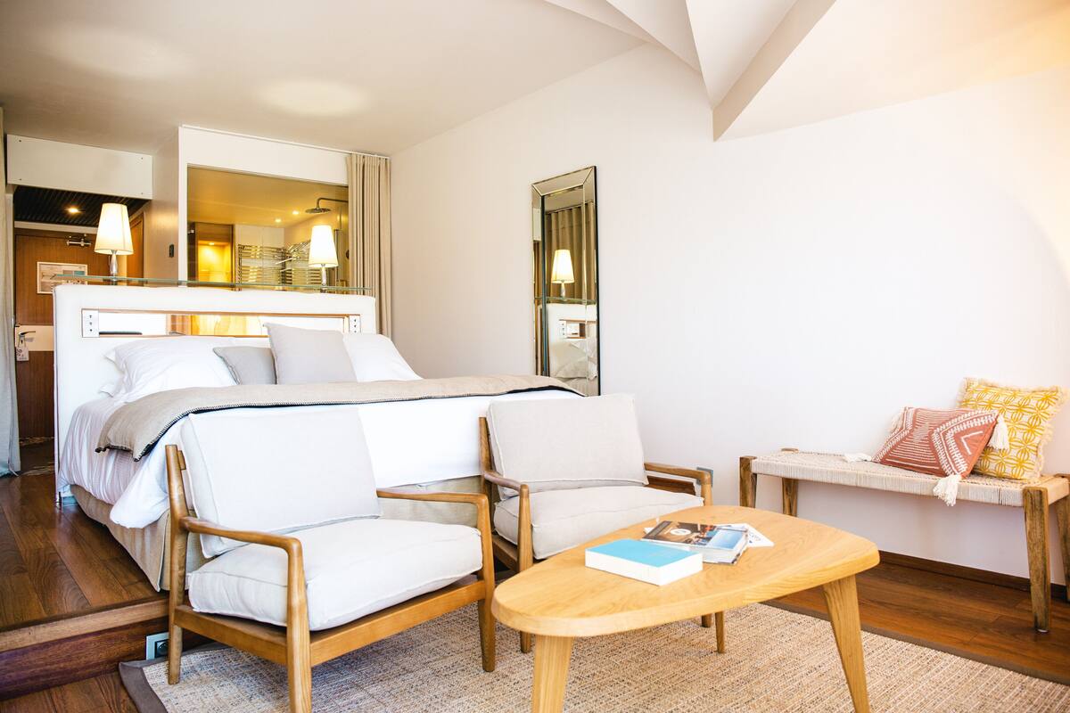 Ein Hotelzimmer mit einem Bett, zwei Sesseln, einem kleinen Tisch, einer Bank und einem Spiegel.