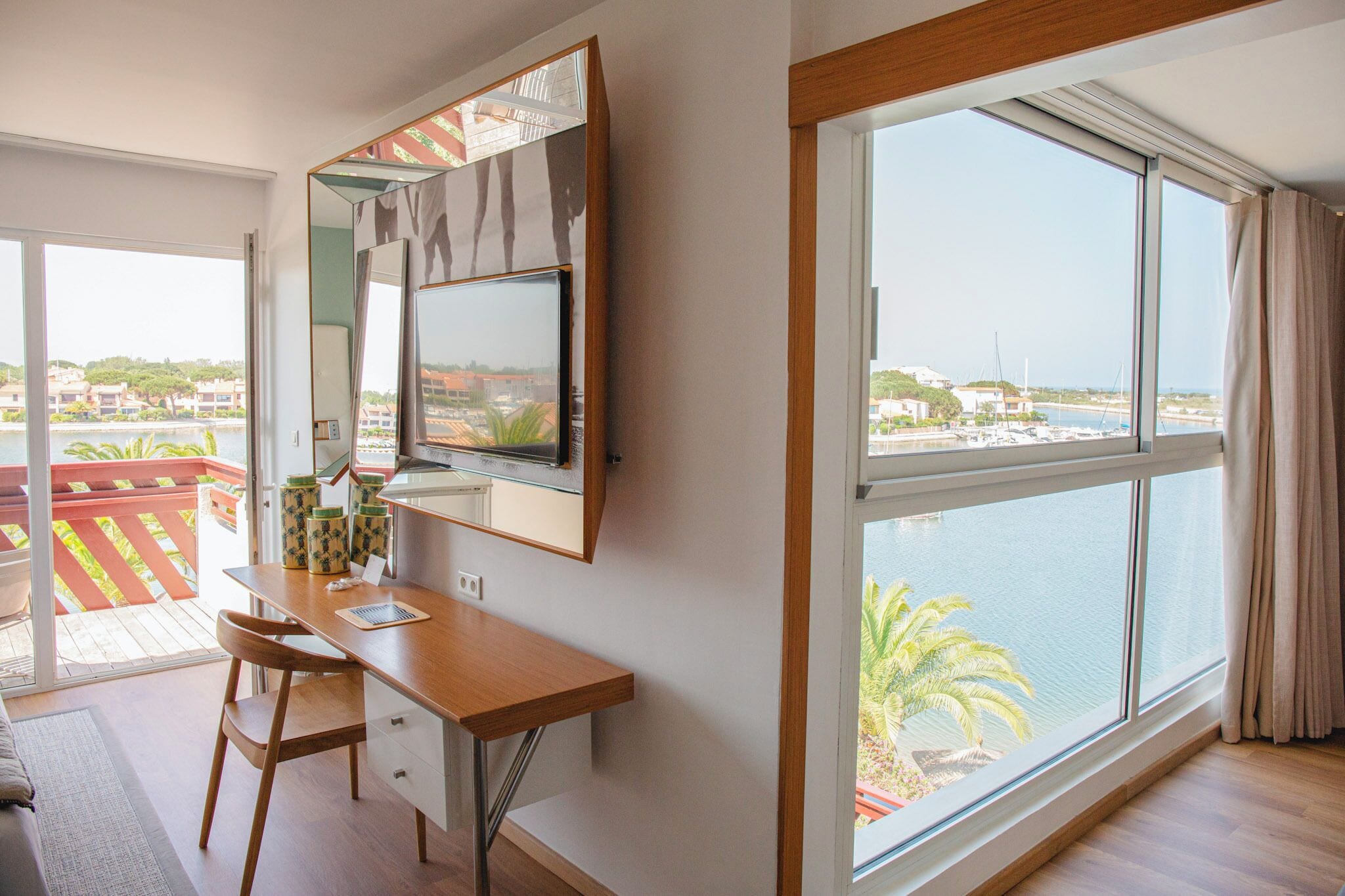 Una habitación con un ventanal grande, un escritorio con una silla, un televisor y vistas a un cuerpo de agua y edificios.