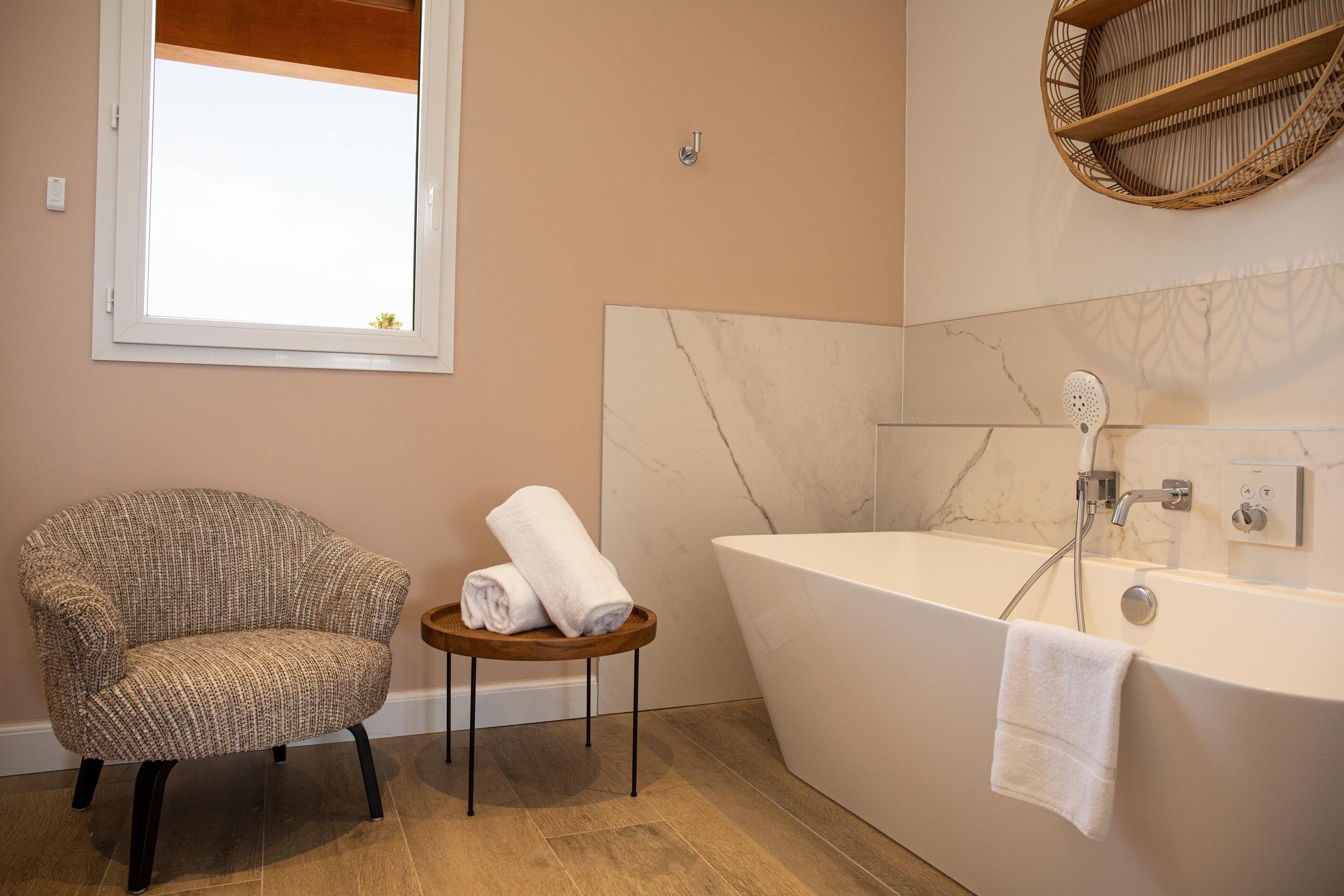A bathroom with a large bathtub, a chair, and a small wooden stool with towels.