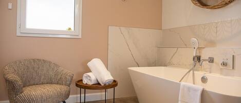 A bathroom with a large bathtub, a chair, and a small wooden stool with towels.