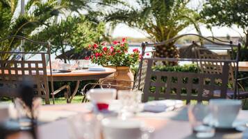 Un espacio al aire libre para comer con una mesa dispuesta para dos personas, rodeada de sillas, una planta en maceta con flores rojas y vistas a palmeras y un cuerpo de agua.