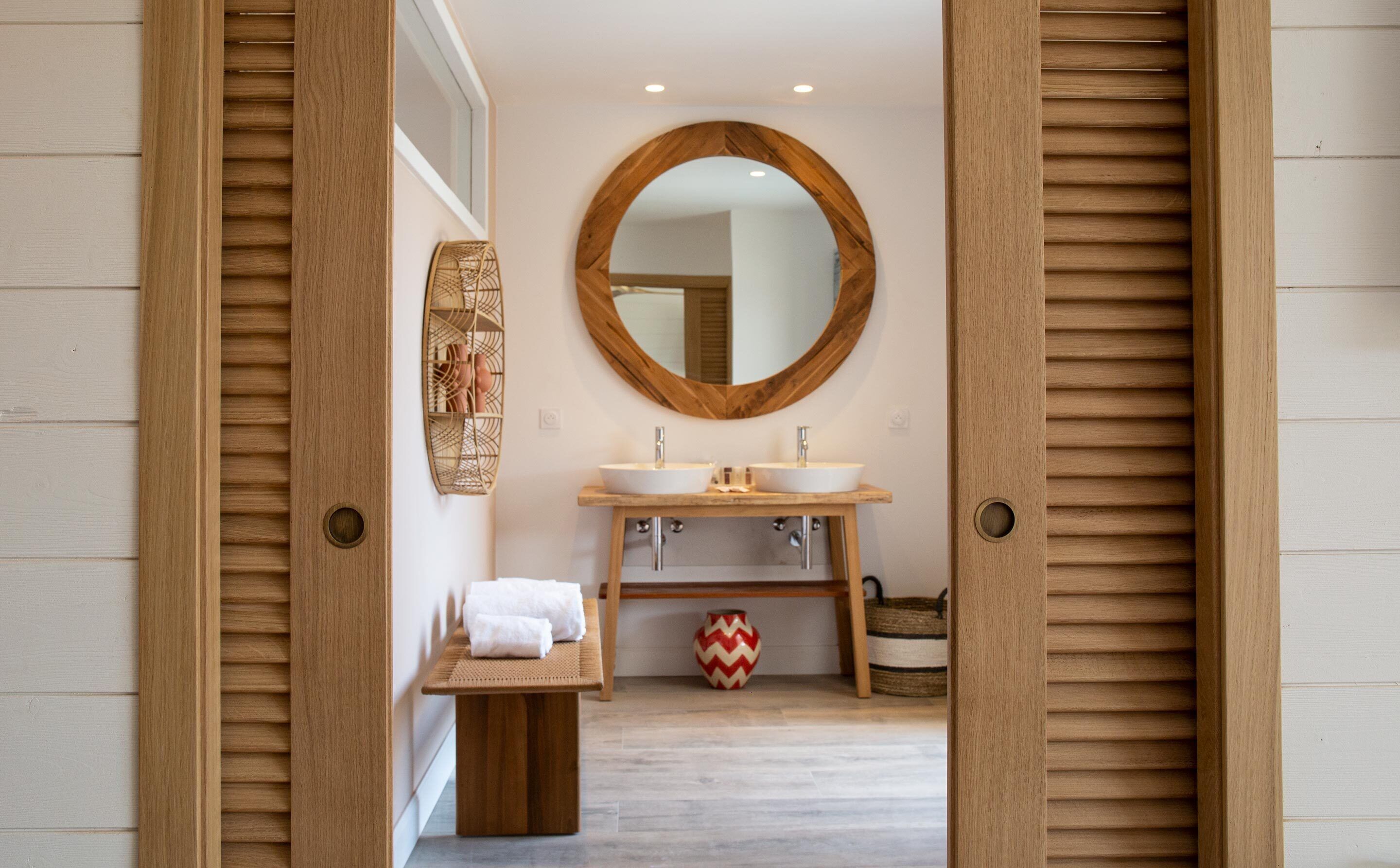 Une salle de bain avec un lambris en bois, un miroir rond, deux lavabos, une petite table avec des serviettes et un vase décoratif.