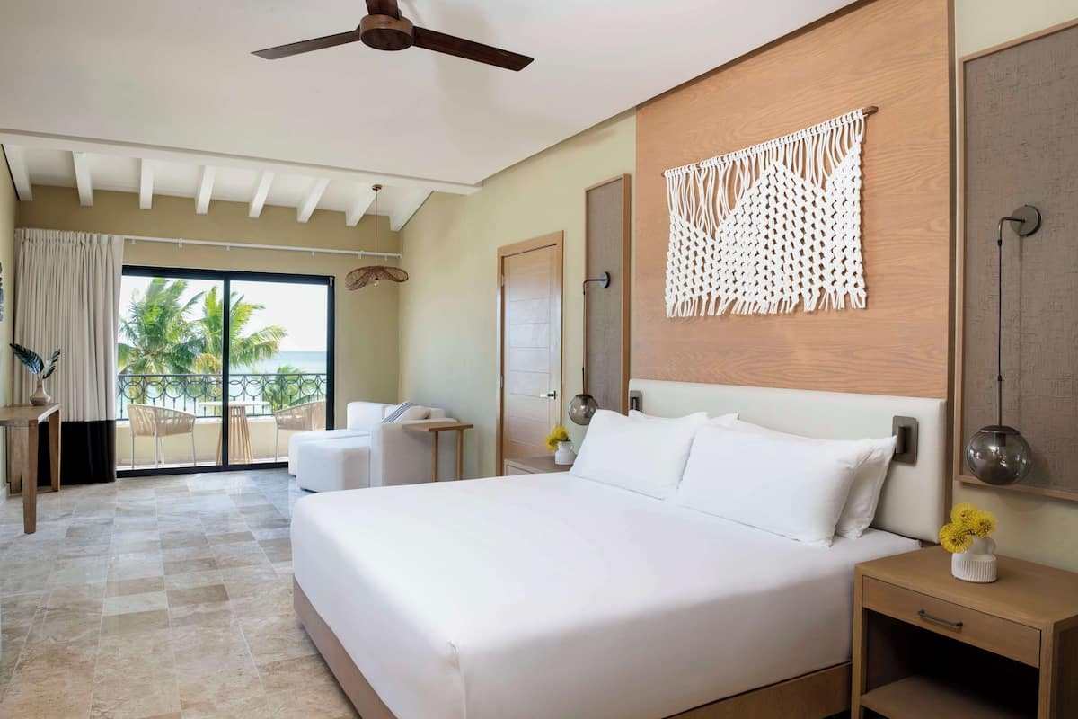 Habitación de hotel con una cama grande, un ventilador de techo, un cabecero de madera, un colgador de macramé en la pared, una mesita de noche con un florero y vistas a palmeras a través de una puerta corrediza de vidrio.