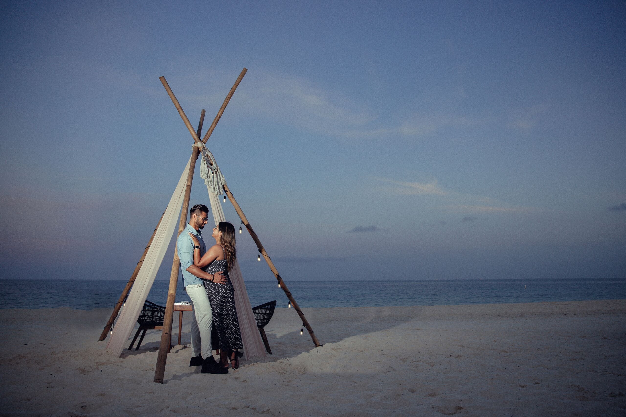A couple sitting under a wooden structure on a beach.