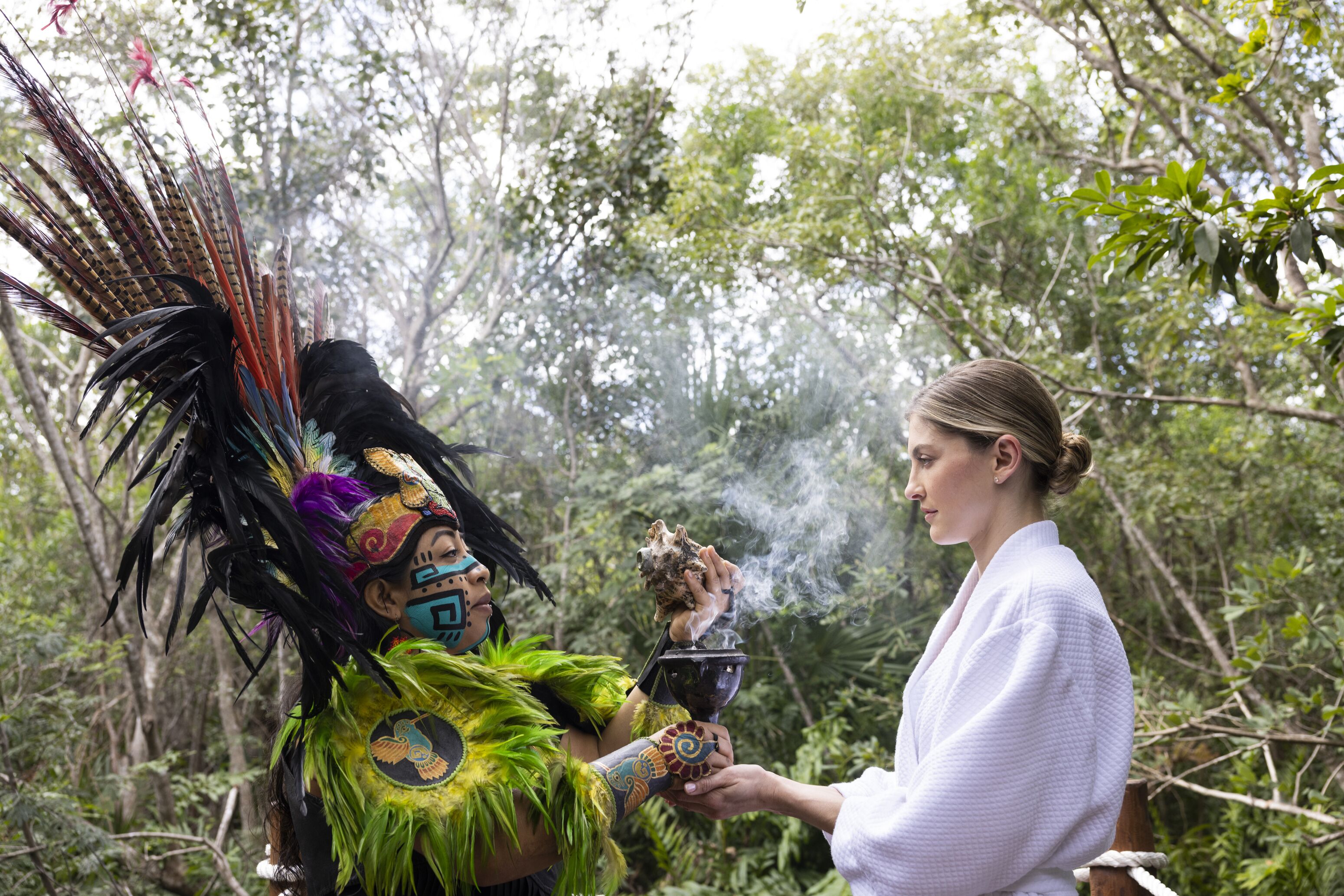Una persona vestida con traje tradicional, tocado por un penacho y un atuendo emplumado, sostiene un recipiente con humo que se eleva, en un entorno natural boscoso.