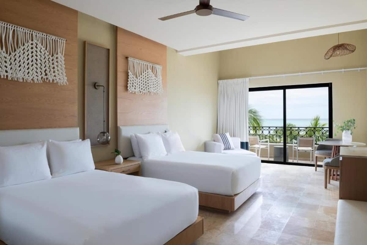Habitación de hotel con dos camas, un balcón con vistas, un ventilador de techo y una pequeña mesa con sillas.