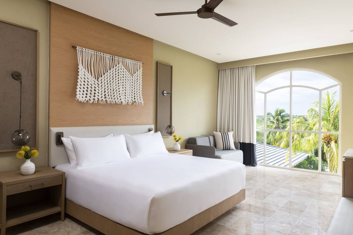 Una cama grande con ropa de cama blanca, un ventilador de techo, un tapiz de macramé, una mesita de noche con un jarrón de flores y vistas a palmeras a través de un ventanal.