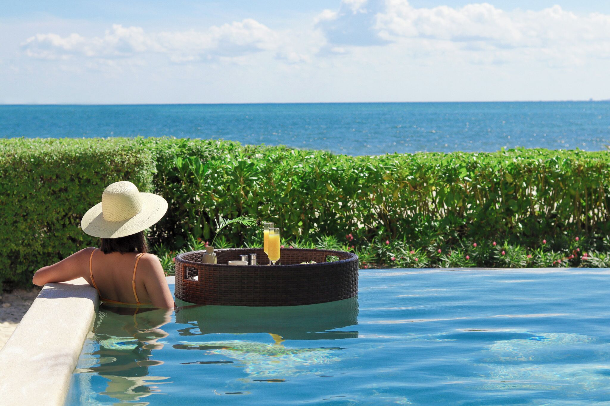 A person relaxing in a pool with a drink and a basket, overlooking the ocean.