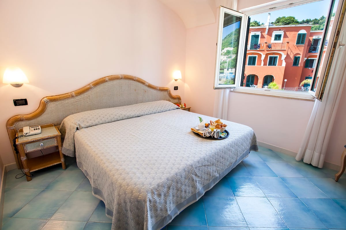 A bedroom with a bed, bedside table, lamp, and a window with a view of buildings.