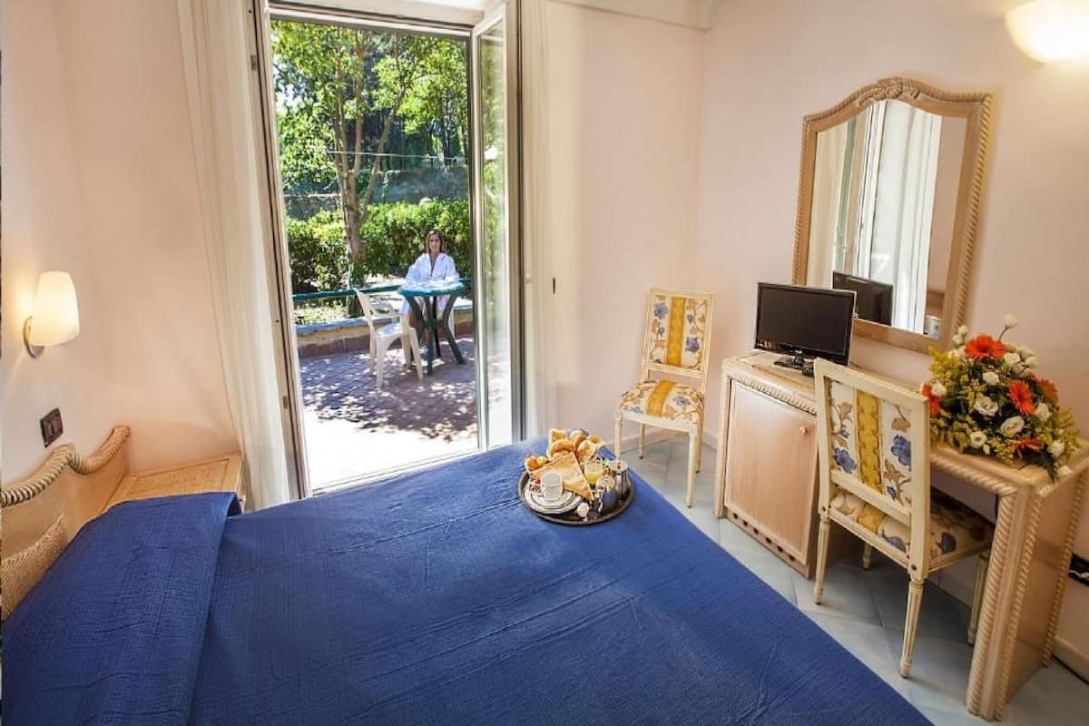 A bedroom with a bed, a desk with a computer, a chair, a mirror, and a view of an outdoor area.