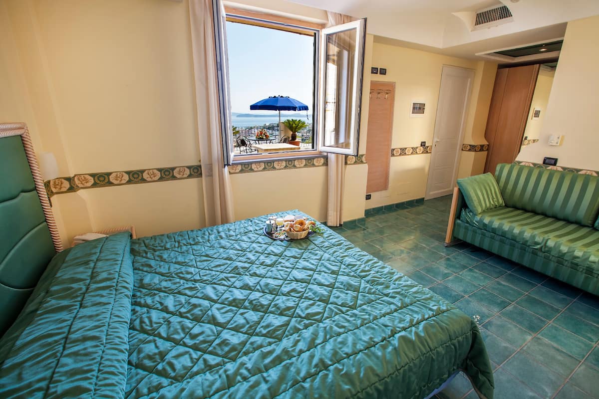 A hotel room with a bed, a sofa, a window with a view of the sea, and a balcony with a table and umbrella.