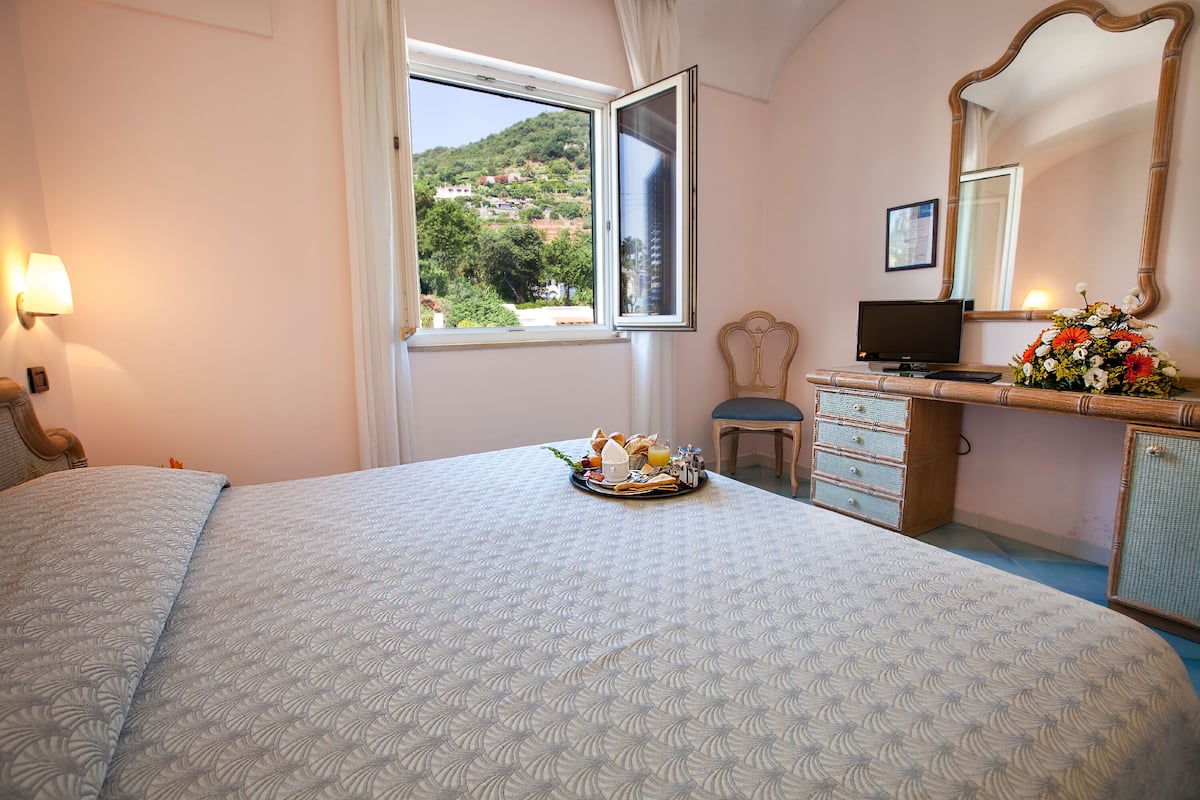 A bedroom with a bed, a desk with a computer, a dresser, a window with a view of greenery, and a mirror.