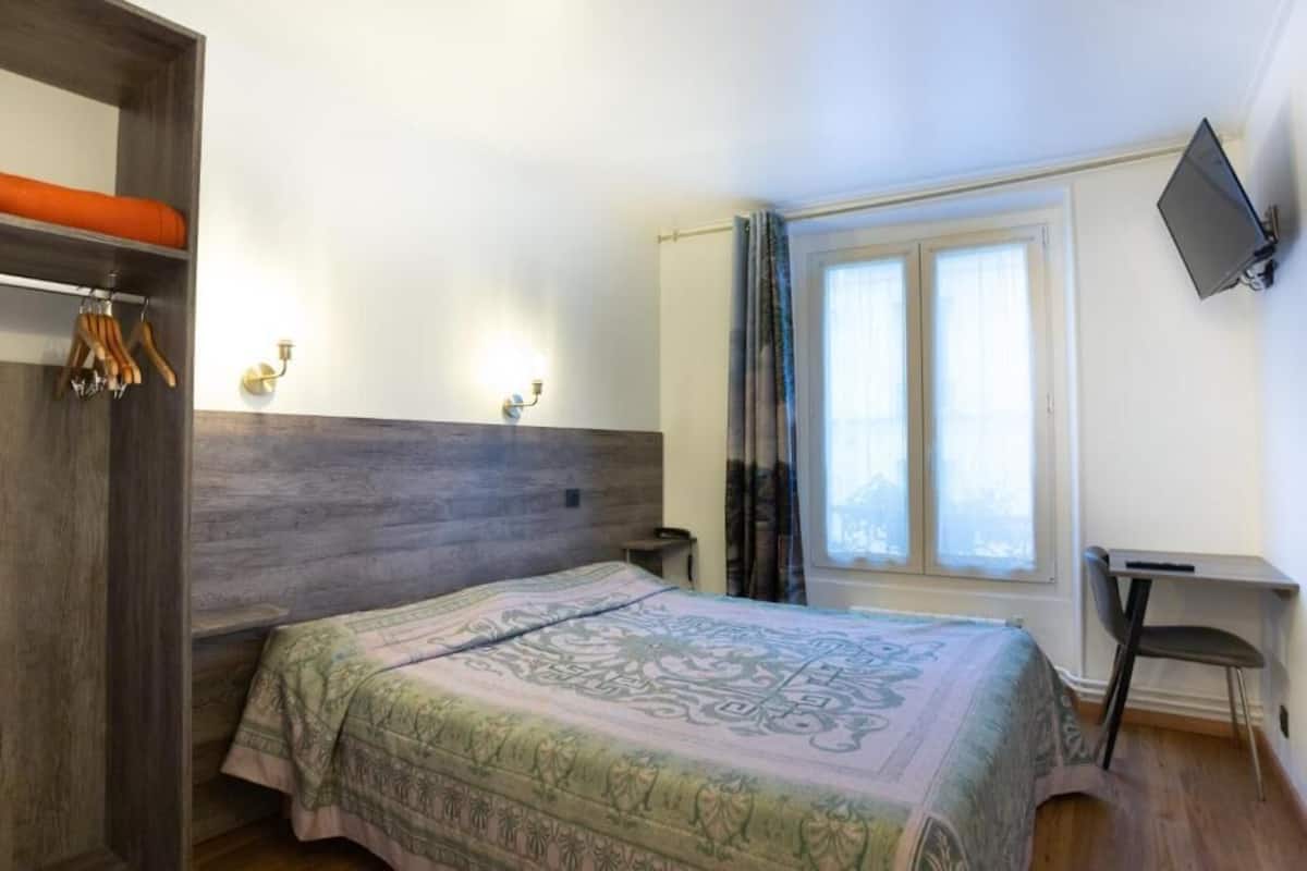 A bedroom with a bed, a desk, a TV, and a window with curtains.
