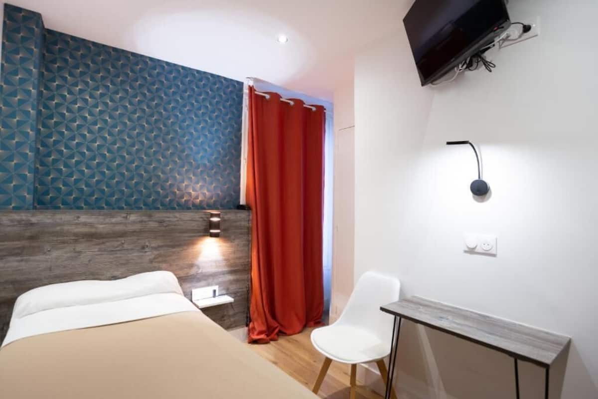 A hotel room with a bed, a desk, a chair, a TV, and a wall with a geometric pattern.