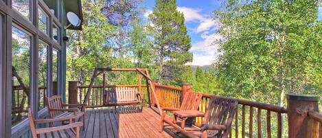 A wooden deck with two wooden chairs and a table, surrounded by a railing, overlooking a forested area.