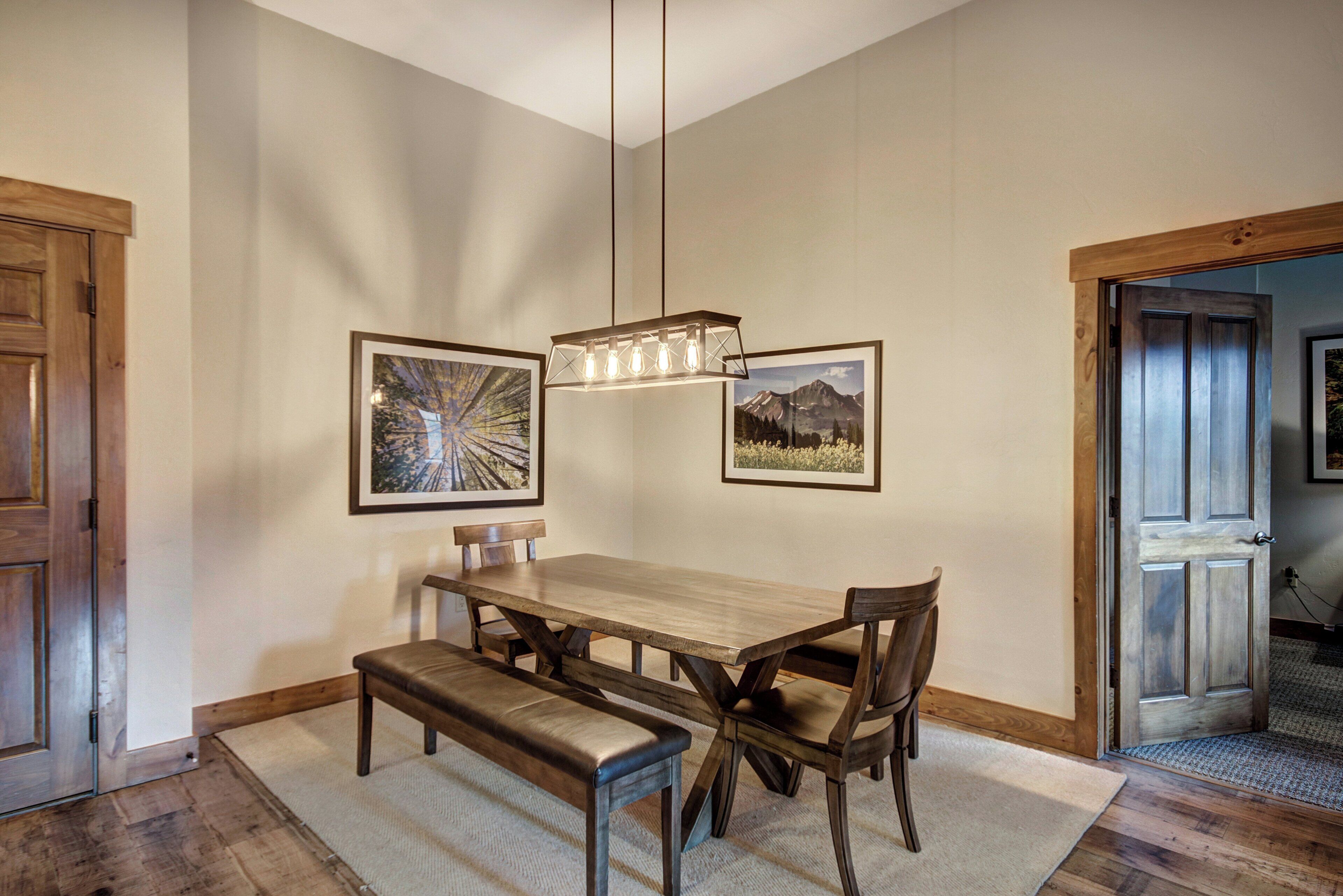A dining area with a wooden table and benches, framed artwork on the walls, and a hanging light fixture.